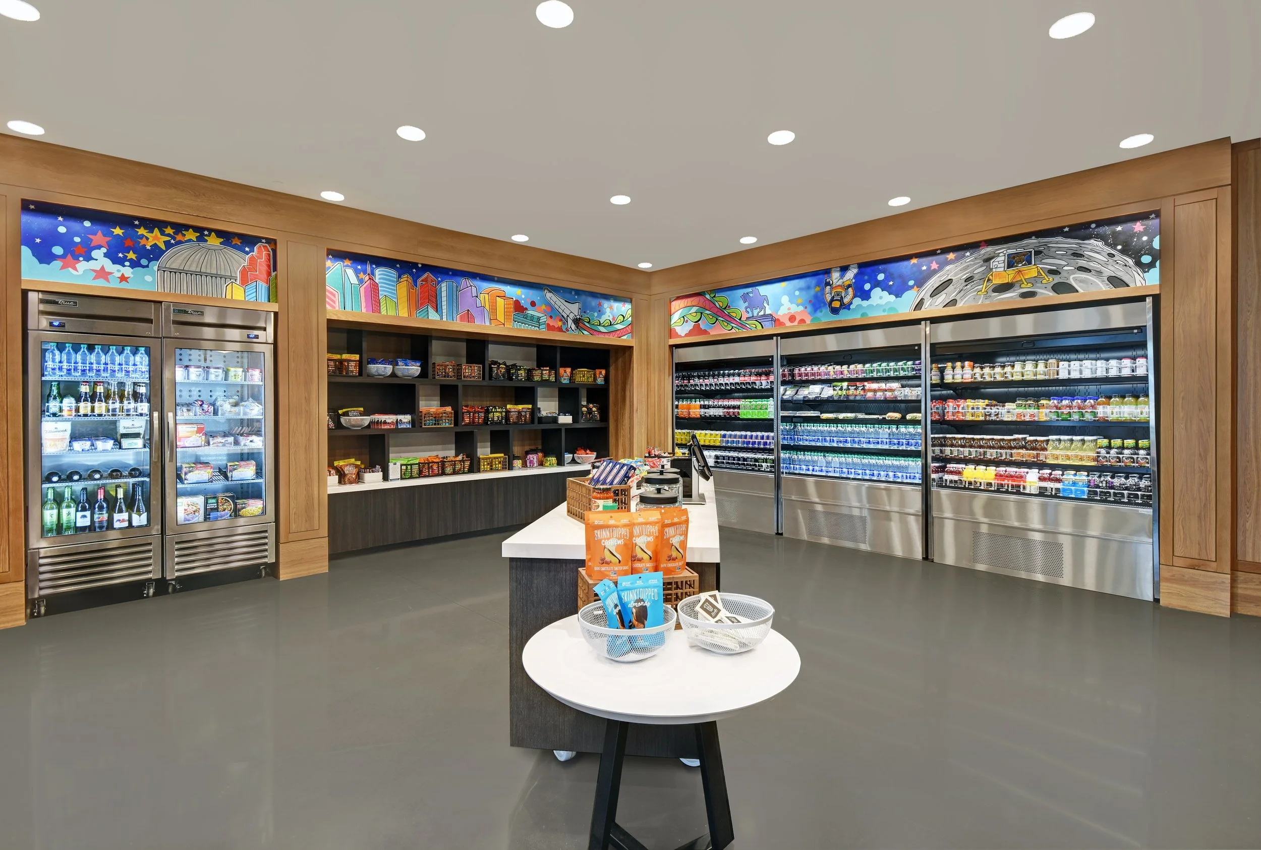 Interior of a convenience store with refrigerated drinks, snacks on shelves, and colorful cityscape and space-themed mural on the walls.