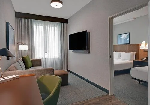 Hotel room with a sofa, green accent pillow, armchair, wall-mounted TV, window with sheer curtains, and a bedroom visible through an open door with a bed and bedside lamps.