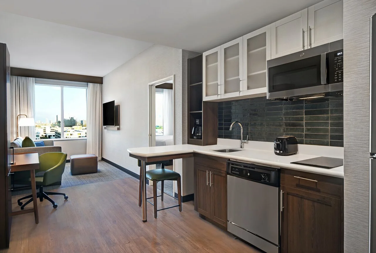 Interior of a modern hotel studio apartment with a kitchen and living area, featuring a window view of a cityscape.