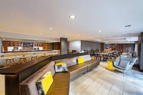 Spacious lounge area with modern furniture, including a long brown sofa with yellow and gray accent pillows, a gray armchair with yellow accent pieces, and a marble counter with barstools, in a well-lit room with tiled flooring and multiple seating a