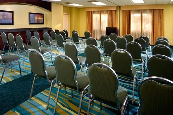 Empty conference room with rows of green chairs, three wall-mounted screens, and large windows with curtains.