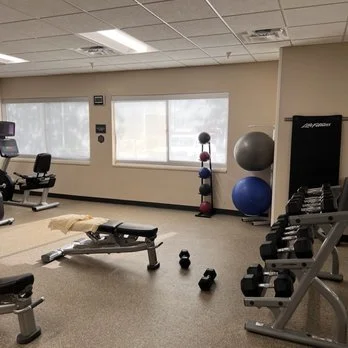 Empty gym with workout equipment, including a bench, dumbbells, kettlebell, and exercise balls near windows.