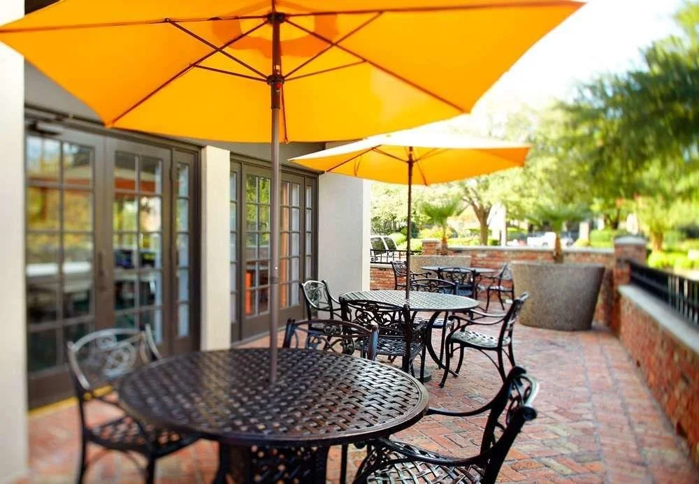 Outdoor patio with metal tables and chairs under large yellow umbrellas, surrounded by brick walls and trees.