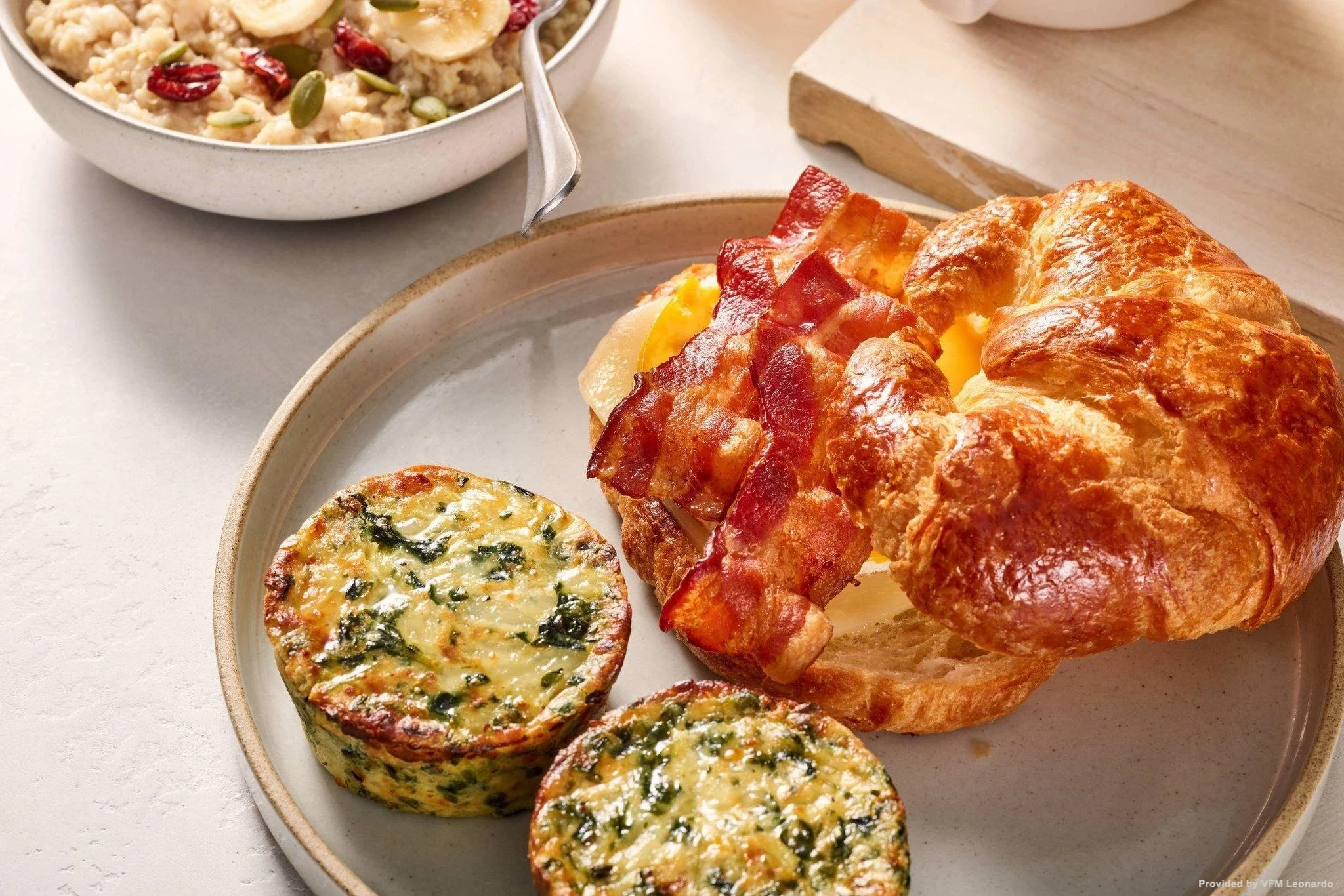 A breakfast plate with a croissant sandwich containing bacon, cheese, and scrambled eggs, served with two spinach and cheese egg muffins, and a bowl of oatmeal topped with banana slices, cranberries, and pumpkin seeds.