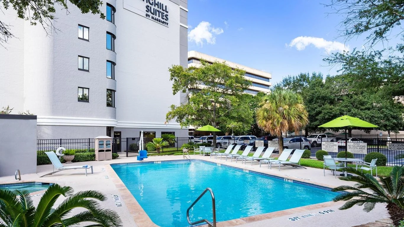 Outdoor hotel pool area with lounge chairs, umbrellas, and tropical plants, surrounded by a black fence and trees, with a tall white hotel building in the background under a partly cloudy sky.