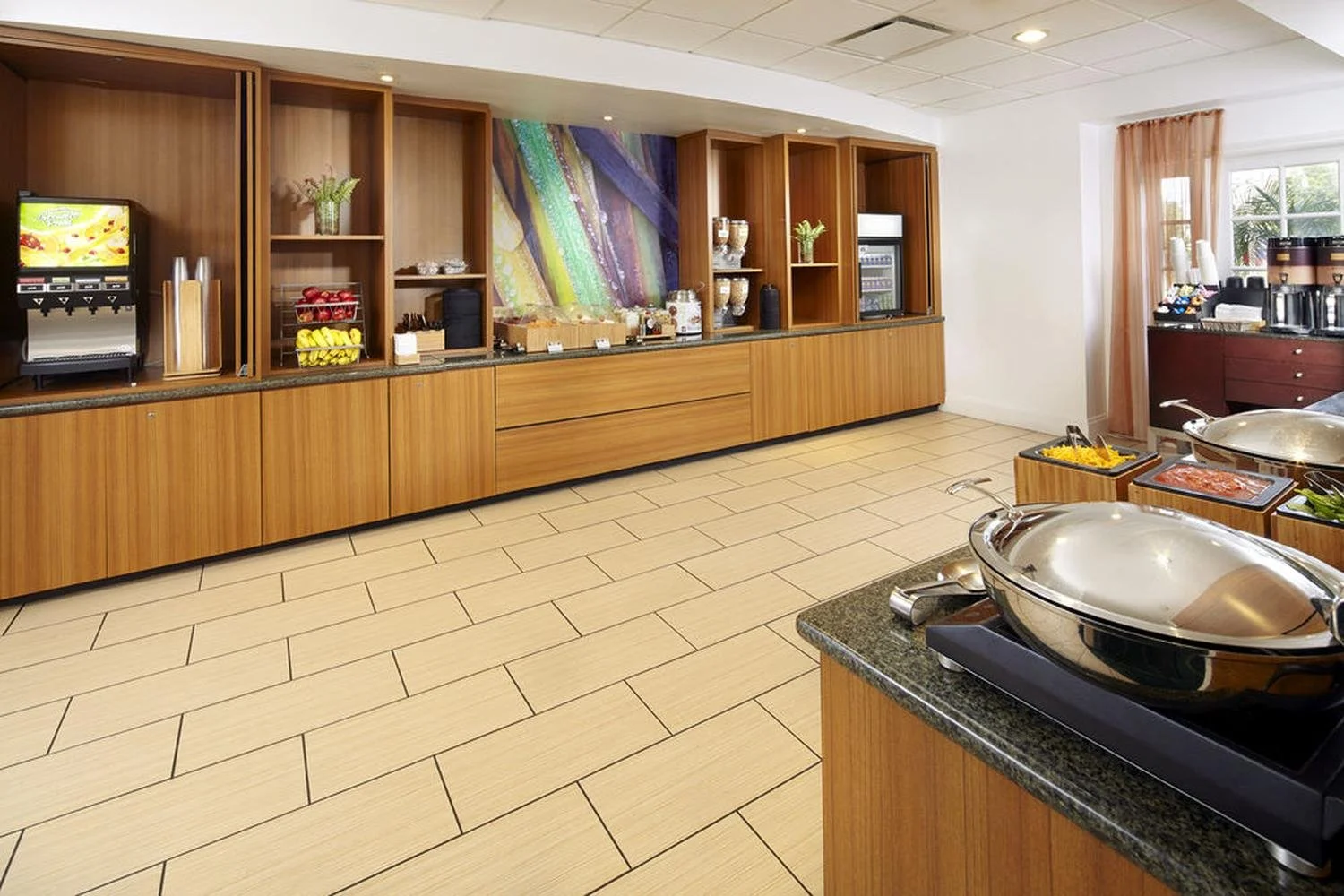 Empty buffet breakfast area with wooden cabinets, countertop, and serving stations, featuring breakfast items, coffee machines, and juice dispensers, with sunlight coming through windows.
