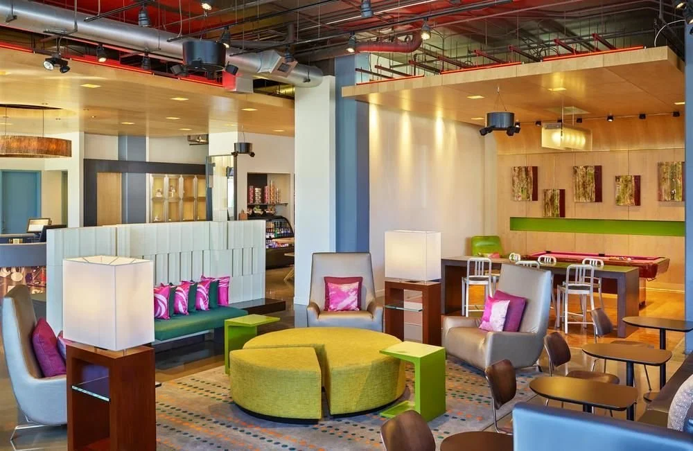 Modern hotel lobby with colorful furniture, including gray armchairs with pink pillows, a green round sofa, and small side tables. There are lamps, artwork on the wall, and a pool table in the background.