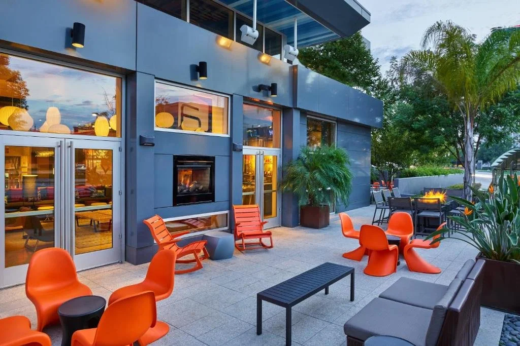 Outdoor patio of a modern building with orange chairs, a black fireplace, tables, potted plants, and trees, during sunset.