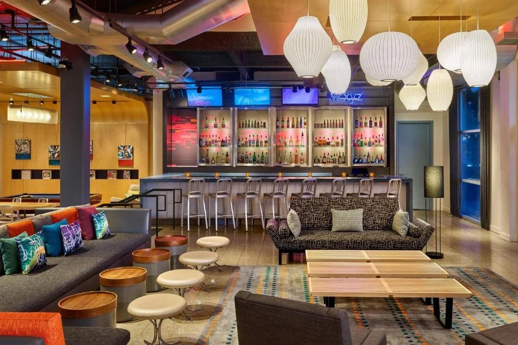 Interior of a modern bar and lounge area with colorful couches, stools, a well-stocked bar with bottles on display, and large white pendant lights hanging from the ceiling.