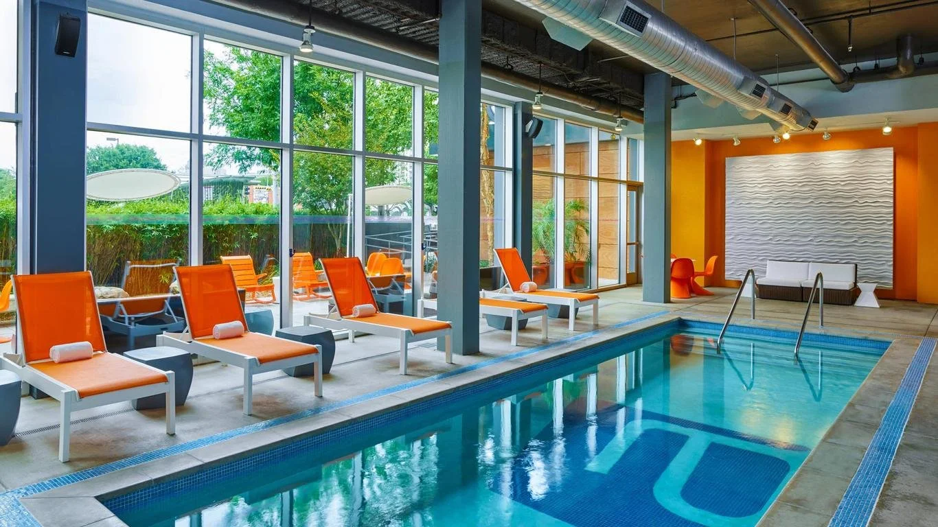 Indoor swimming pool area with orange lounge chairs along the side, large windows showing outdoor greenery, and a seating area with white and orange chairs at the far end.