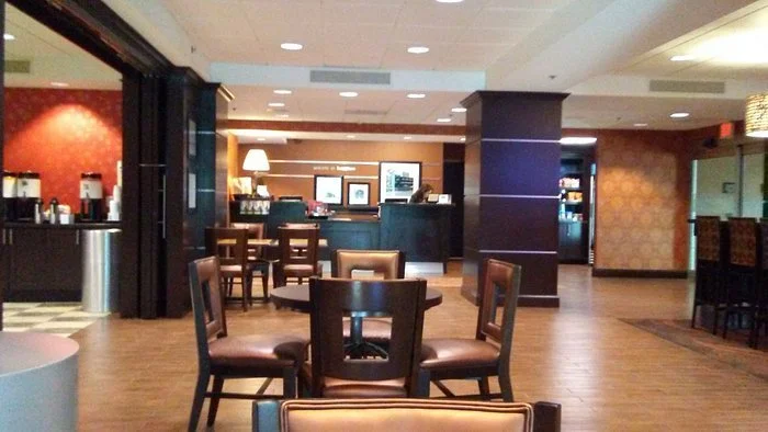 Empty restaurant or cafe with wooden tables and chairs, a counter area with screens, and ambient lighting.