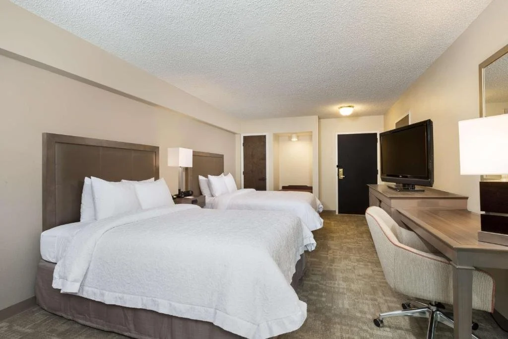 Two queen beds with white bedding in a hotel room, a wall-mounted TV on a desk, and a chair.