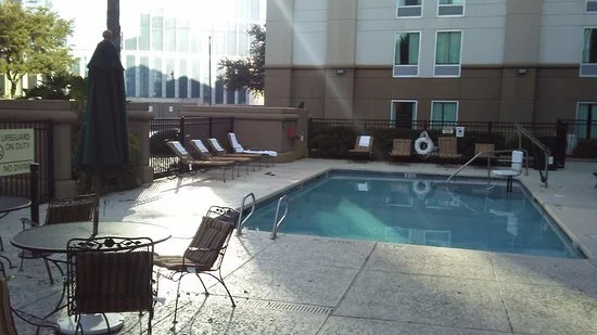 Empty outdoor swimming pool area with chairs, tables, and a building in the background.