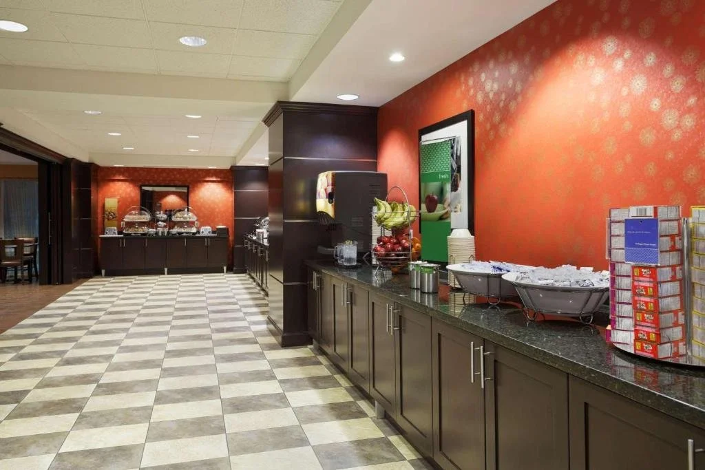 Buffet area in a hotel breakfast lounge with fruit, cereal, and juice containers, featuring dark wood cabinets, checkered floor tiles, with a red textured wall and framed artwork.
