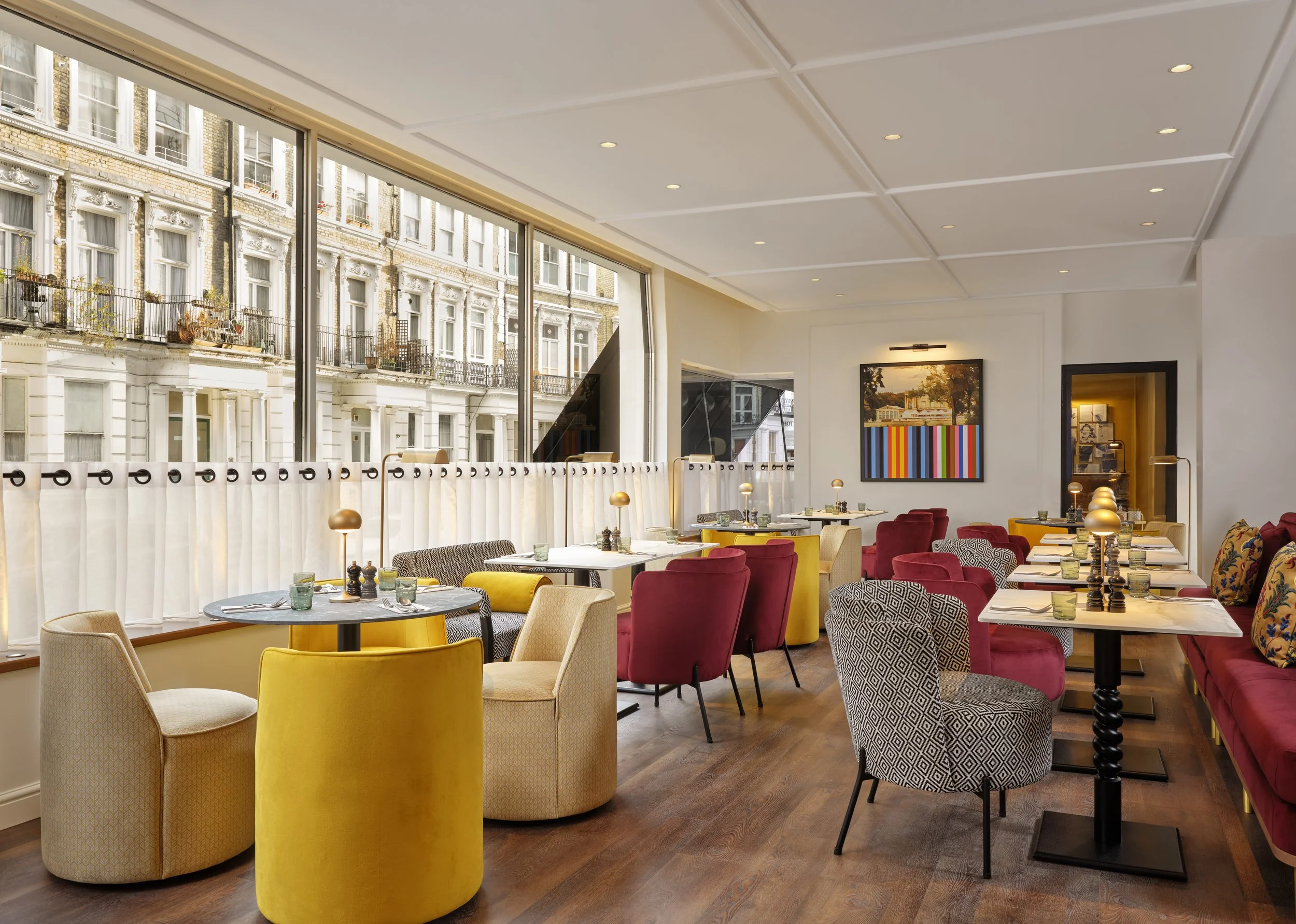 Interior of a modern restaurant or cafe with colorful chairs, tables set with glasses and cutlery, large windows showing historic brick buildings outside, and contemporary artwork on the walls.