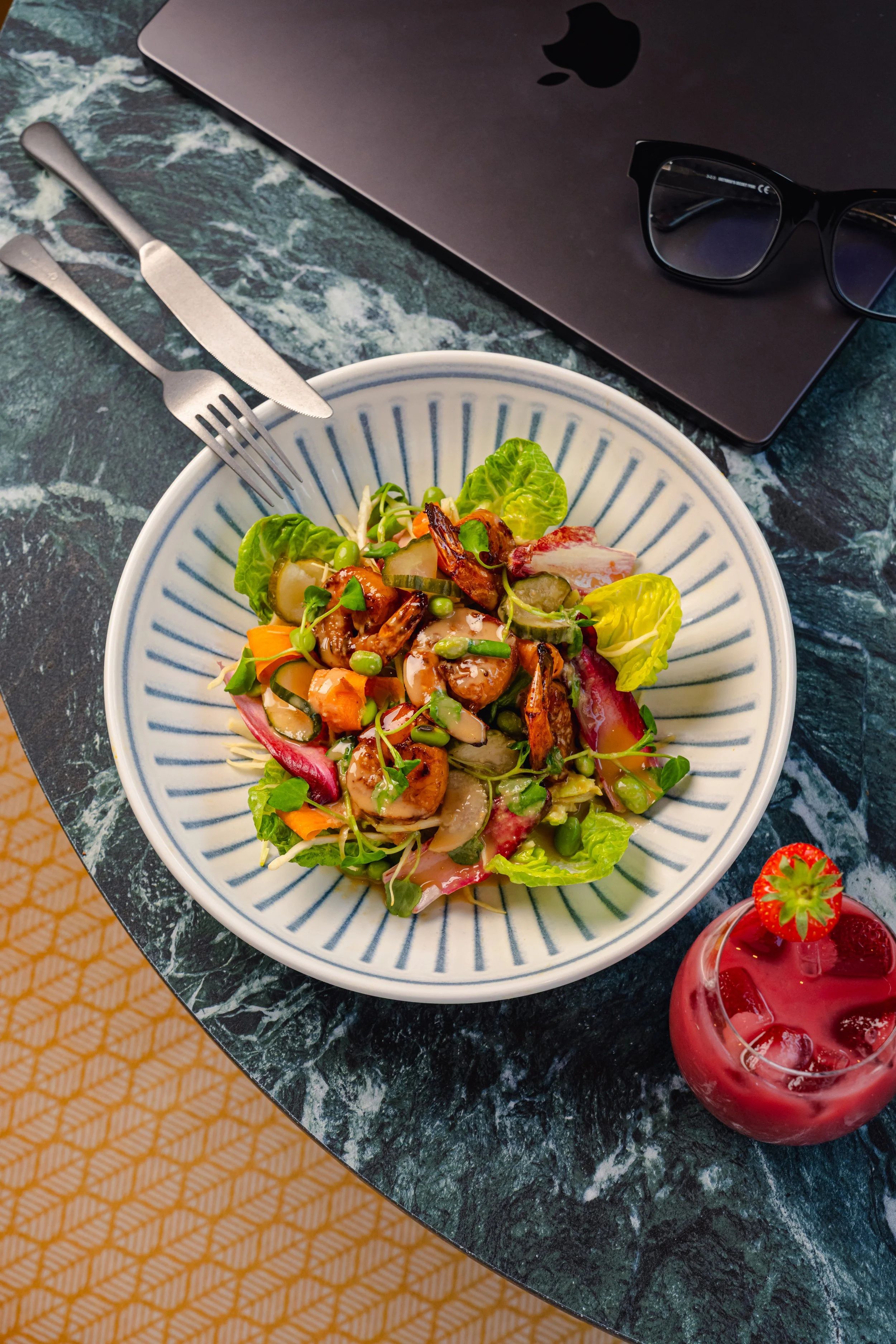 A bowl of salad with mixed greens, cherry tomatoes, peas, and grilled shrimp on a dark green marble table next to an open MacBook laptop, black glasses, and a strawberry-flavored drink with strawberries inside.