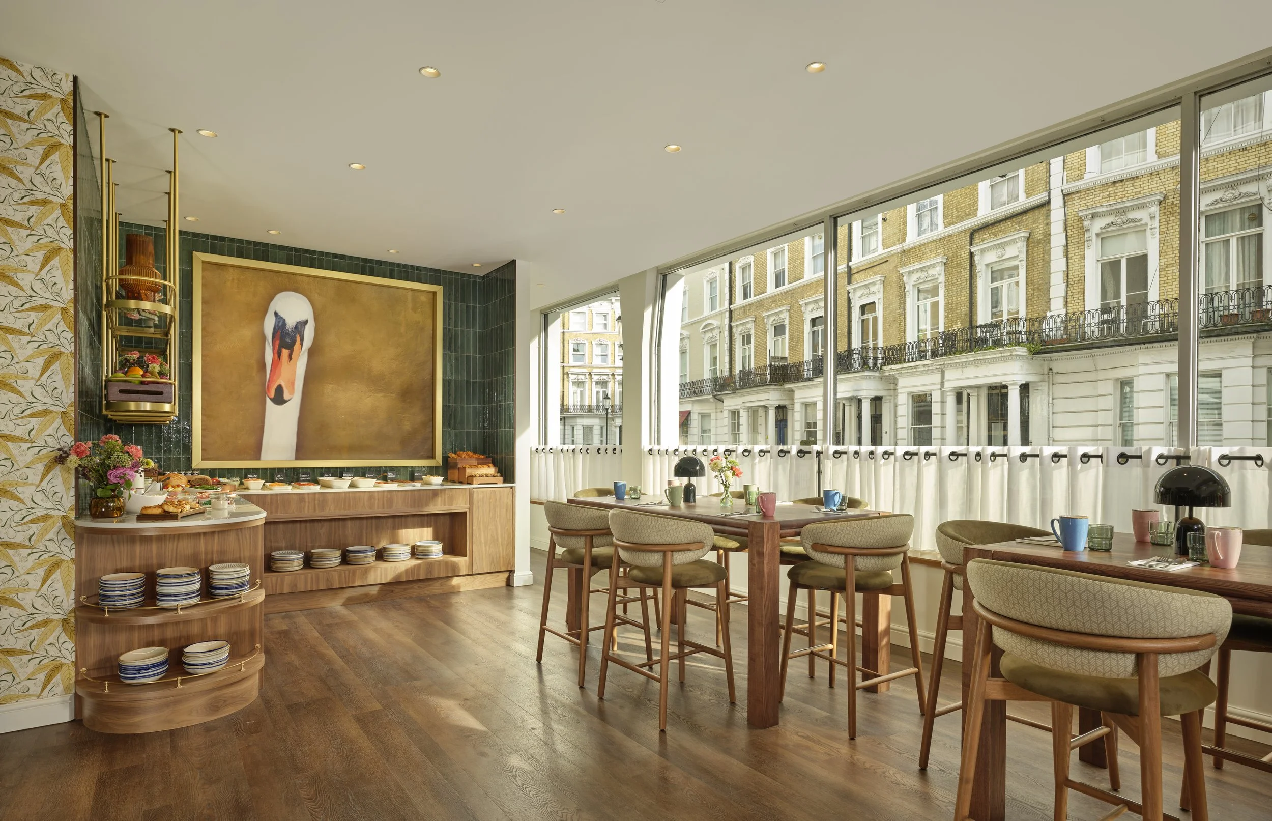 A bright dining area with large windows showing a city street with historic buildings. The room has wooden floors, a buffet with a painting of a swan's head, and tables set with colorful mugs and flowers.