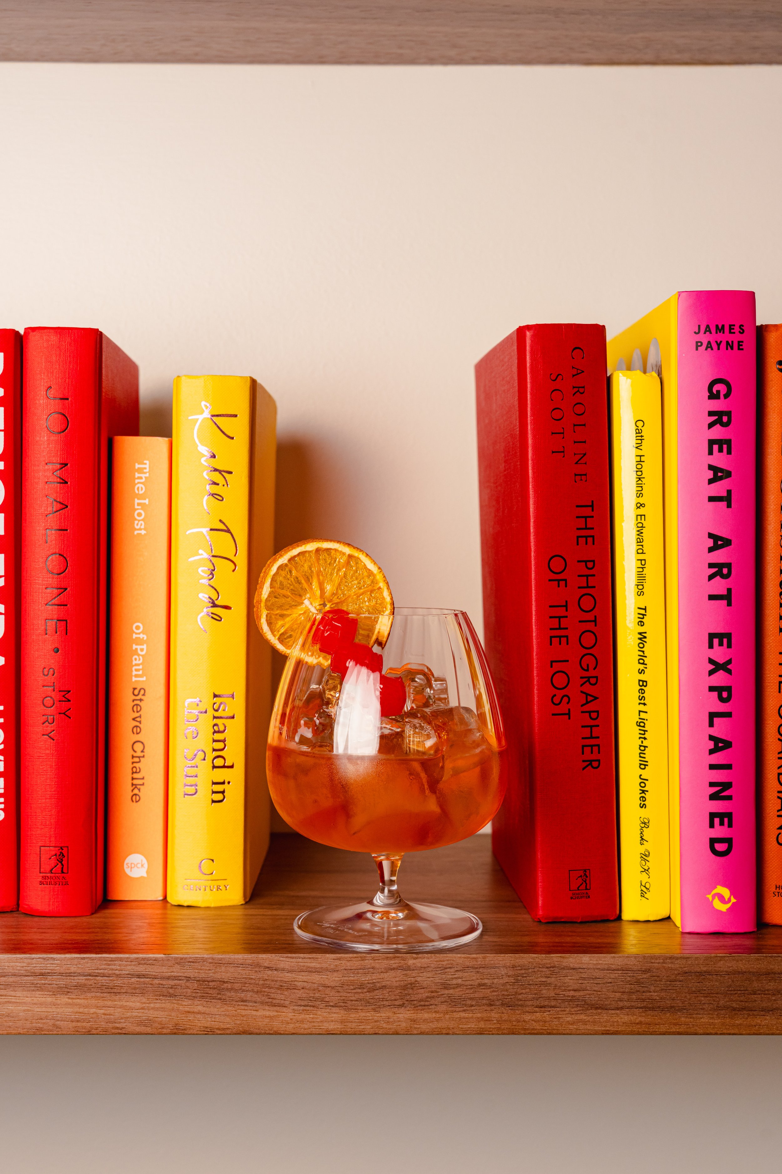 A glass of iced tea with an orange slice garnish in front of a row of colorful books on a wooden shelf.