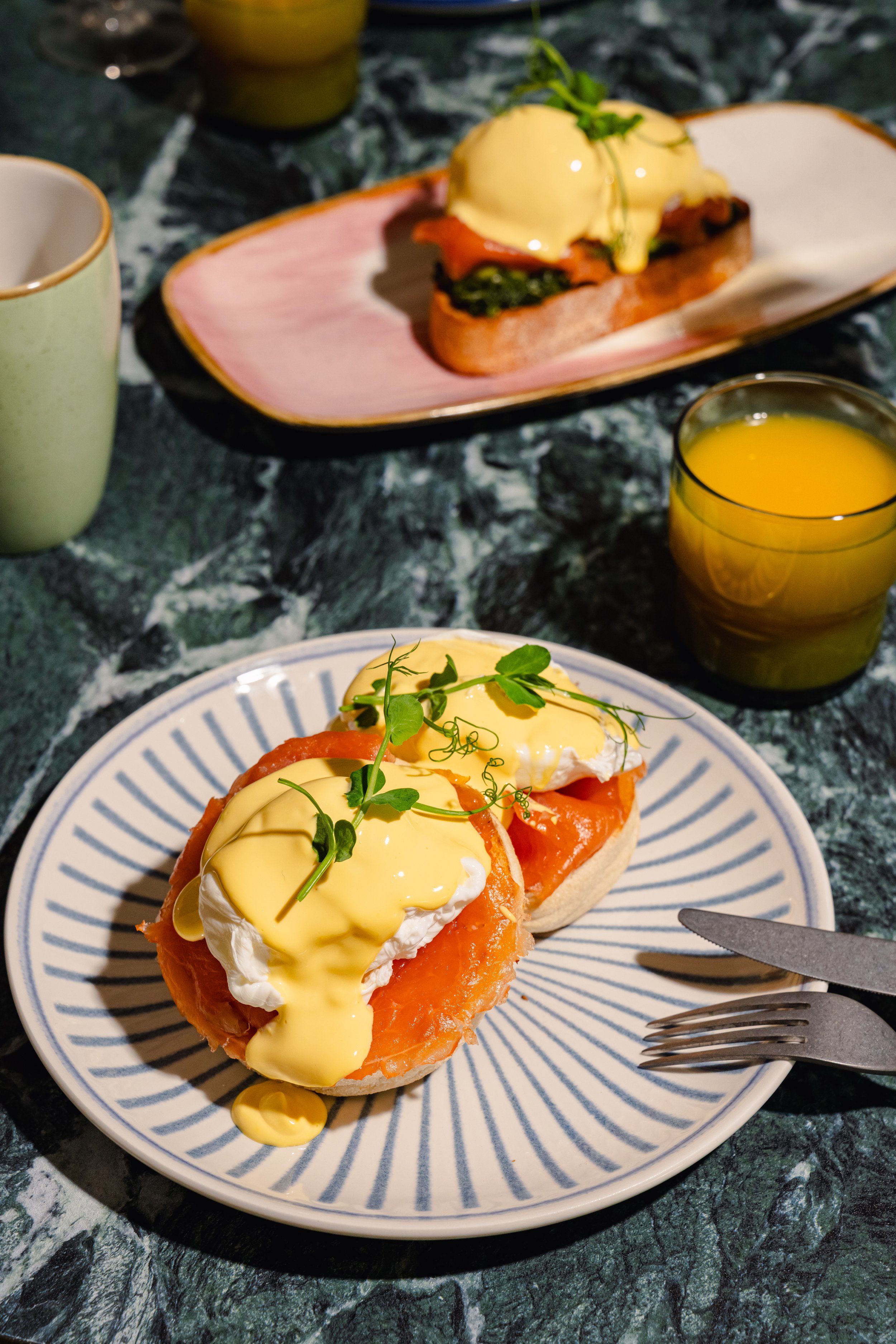 Poached eggs with hollandaise sauce on smoked salmon atop a toasted bagel, garnished with microgreens, served on a white plate with blue stripes on a dark marble table.