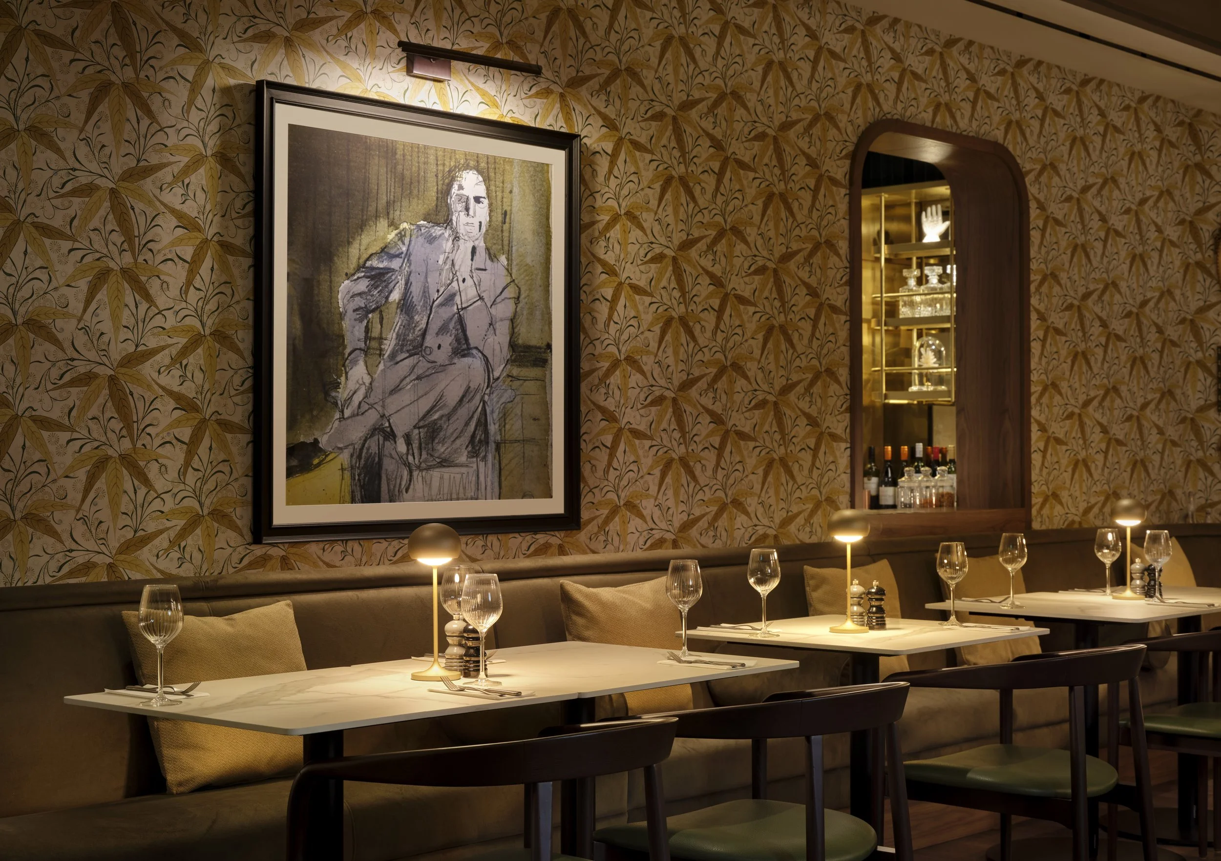Interior of a restaurant dining area with a wall decorated in tropical leaf patterned wallpaper, a framed portrait of a man in a gray suit, and tables set for dining with wine glasses, candles, and napkins.