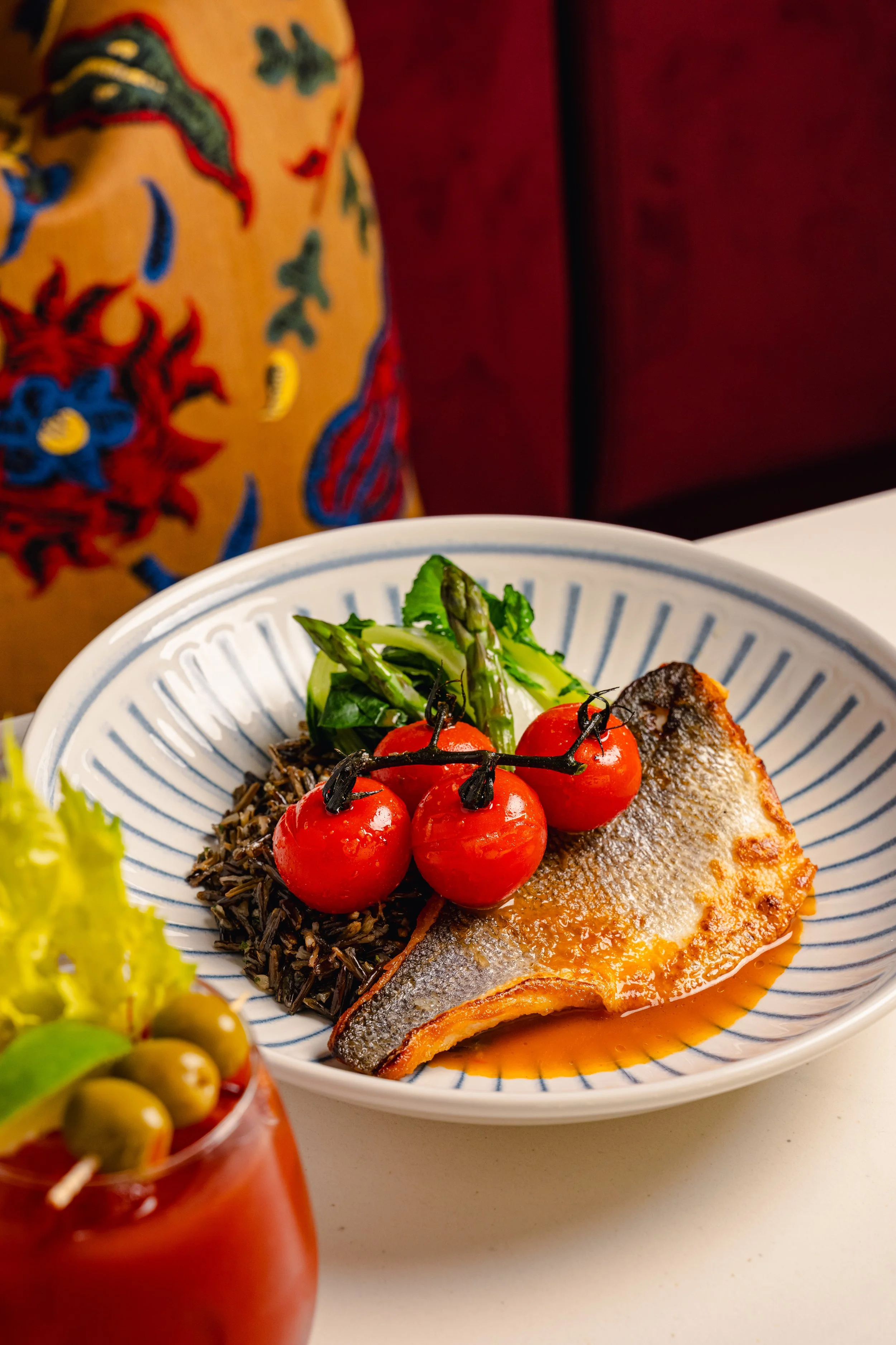 A bowl of grilled fish served with cherry tomatoes, green asparagus, and rice. A red drink with olives and a skewer is visible in the foreground, and an embroidered fabric with a floral pattern is in the background.