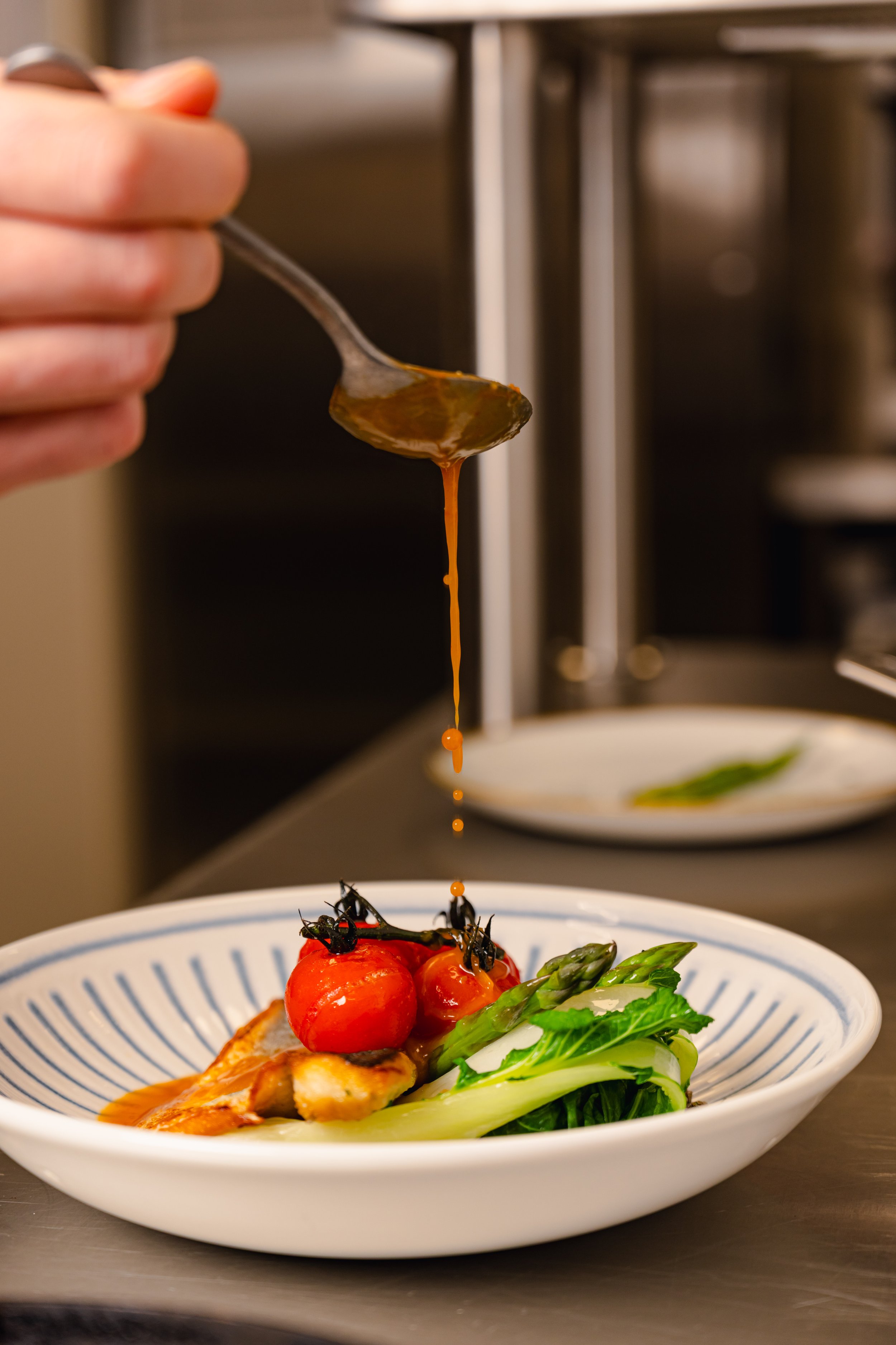 Close-up of a hand pouring sauce or dressing over a plated dish with cherry tomatoes, grilled chicken, and greens.