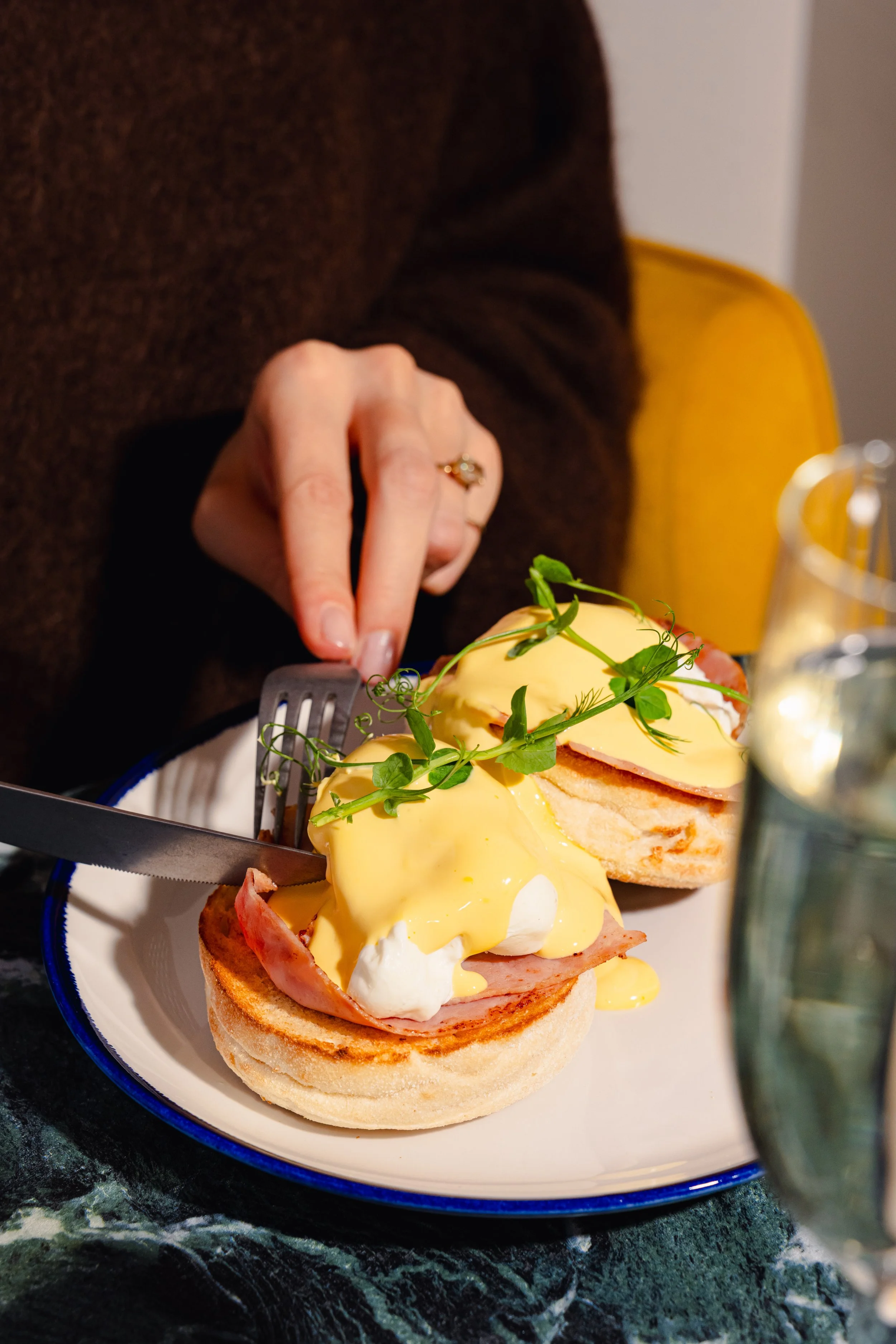 Close-up of an egg's benedict with ham, cheese, and greens on a toasted English muffin, served on a white plate with a blue rim.