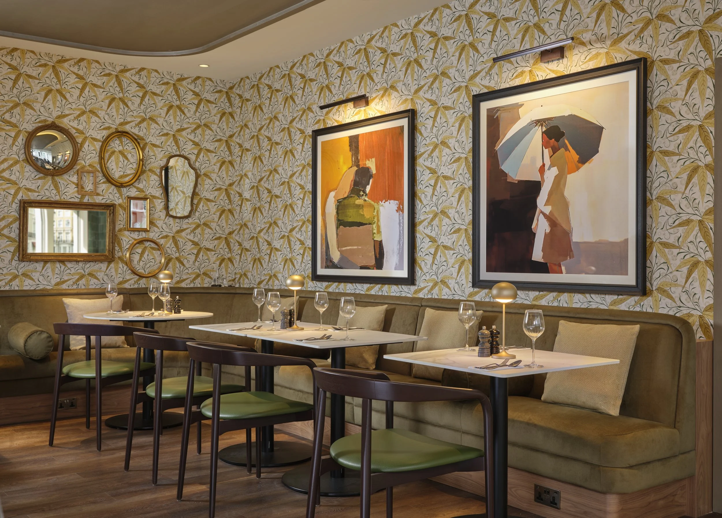 Elegant restaurant interior with wall art and patterned wallpaper, tables set with wine glasses and light fixtures.