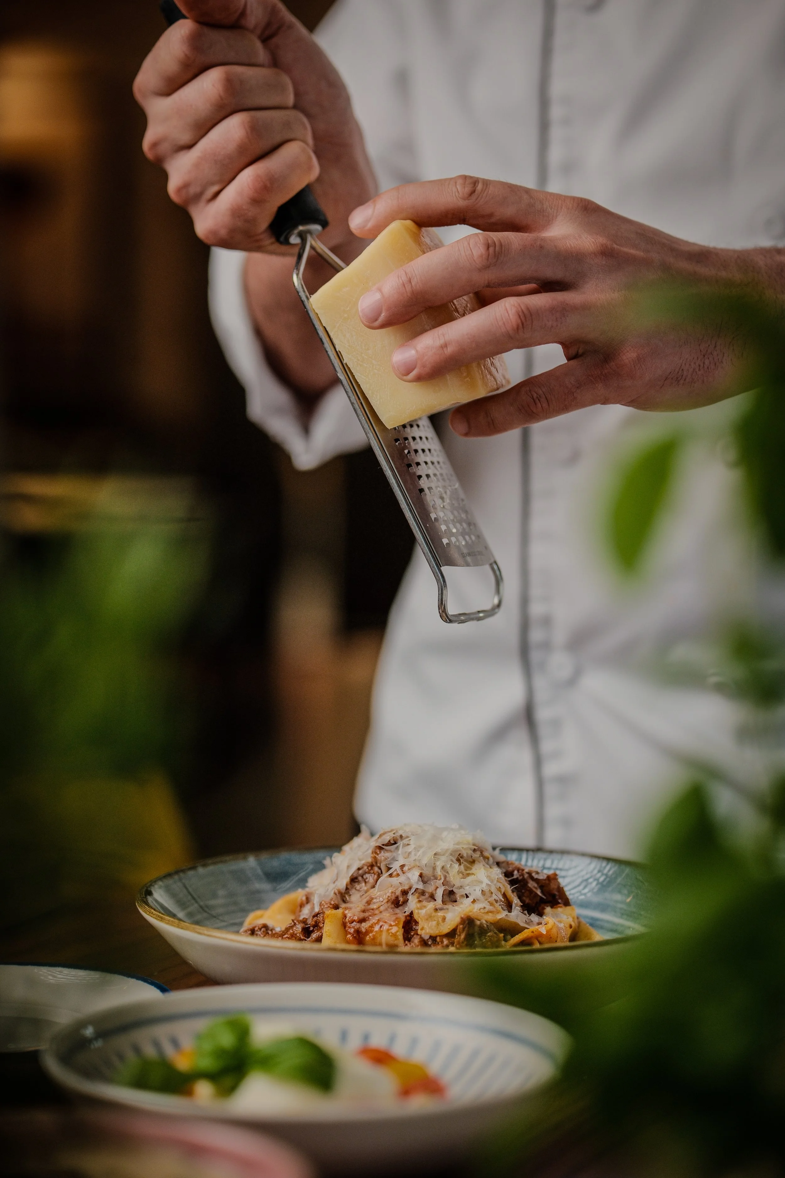 A person grating cheese over a bowl of cooked pasta with meat and vegetables on a dining table.