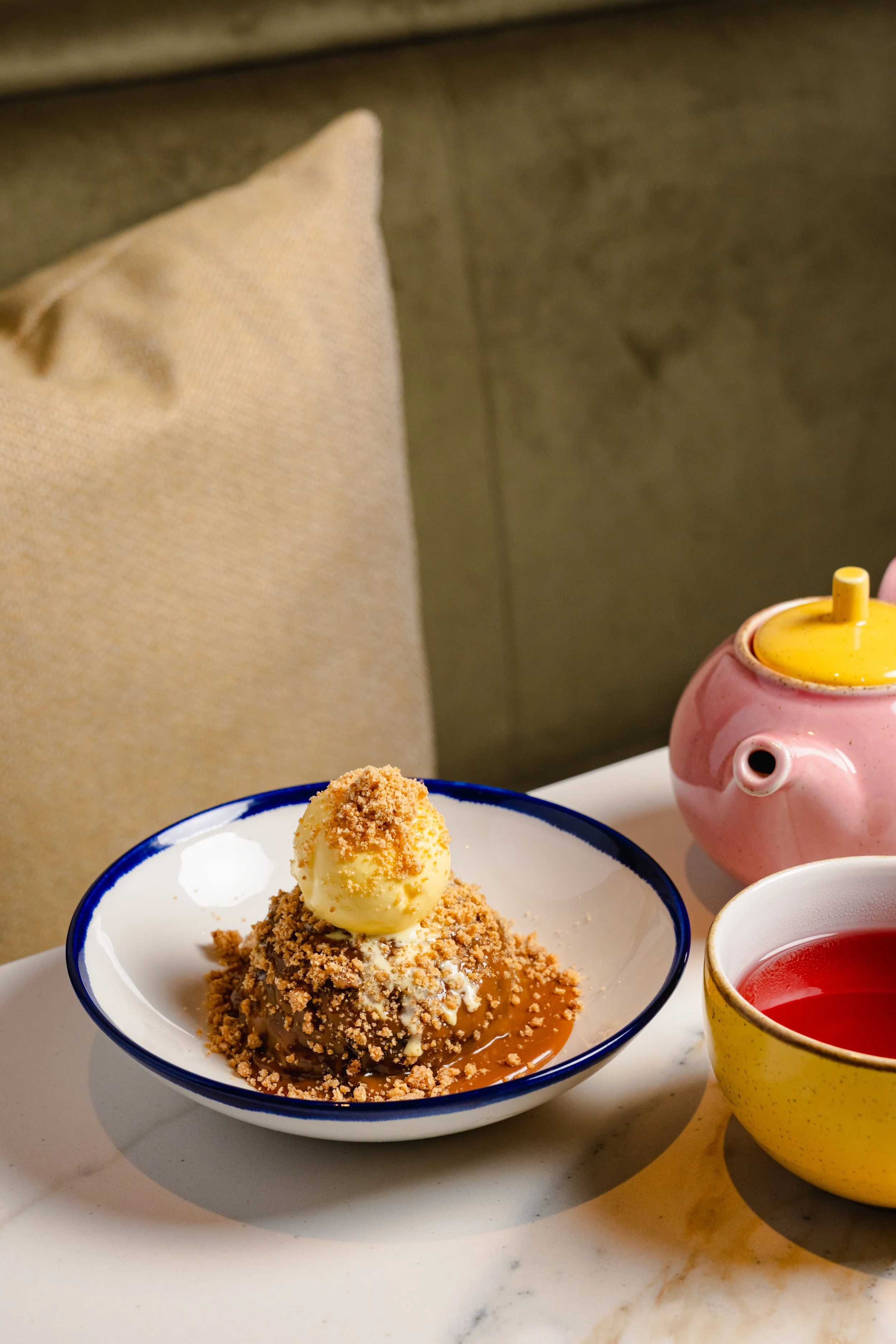 A dessert with a scoop of vanilla ice cream topped with crumbled cookies or nuts, served in a white bowl with a blue rim on a white plate. Next to it is a yellow cup with a pink design filled with red liquid, and a small pink teapot with a yellow lid