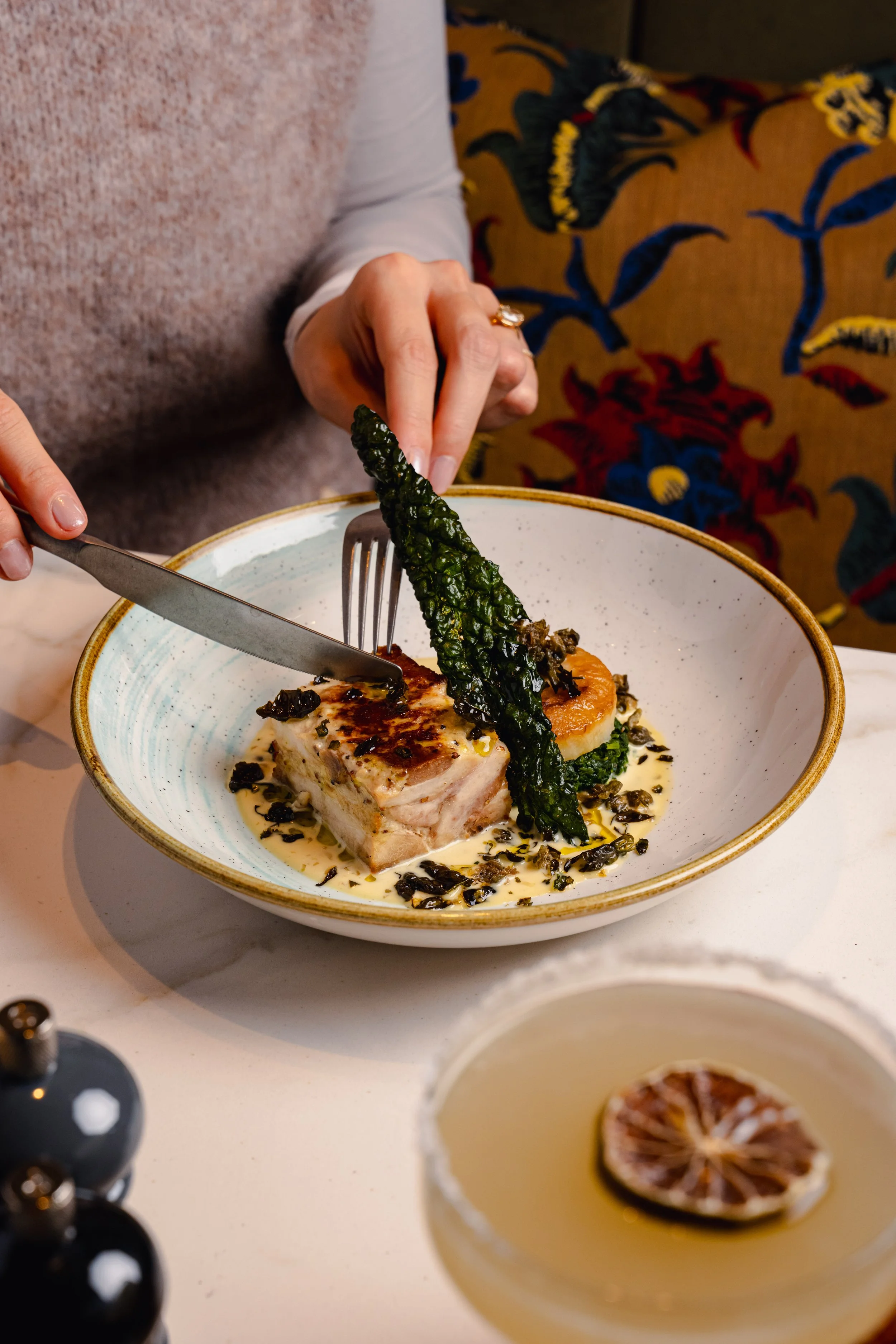 A person is holding a fork and knife near a plated dish that features a piece of cooked meat topped with a glaze, accompanied by a green vegetable, on a light-colored plate. A beverage with a slice of lemon is in the foreground, and a cloth with a colorful pattern is in the background.