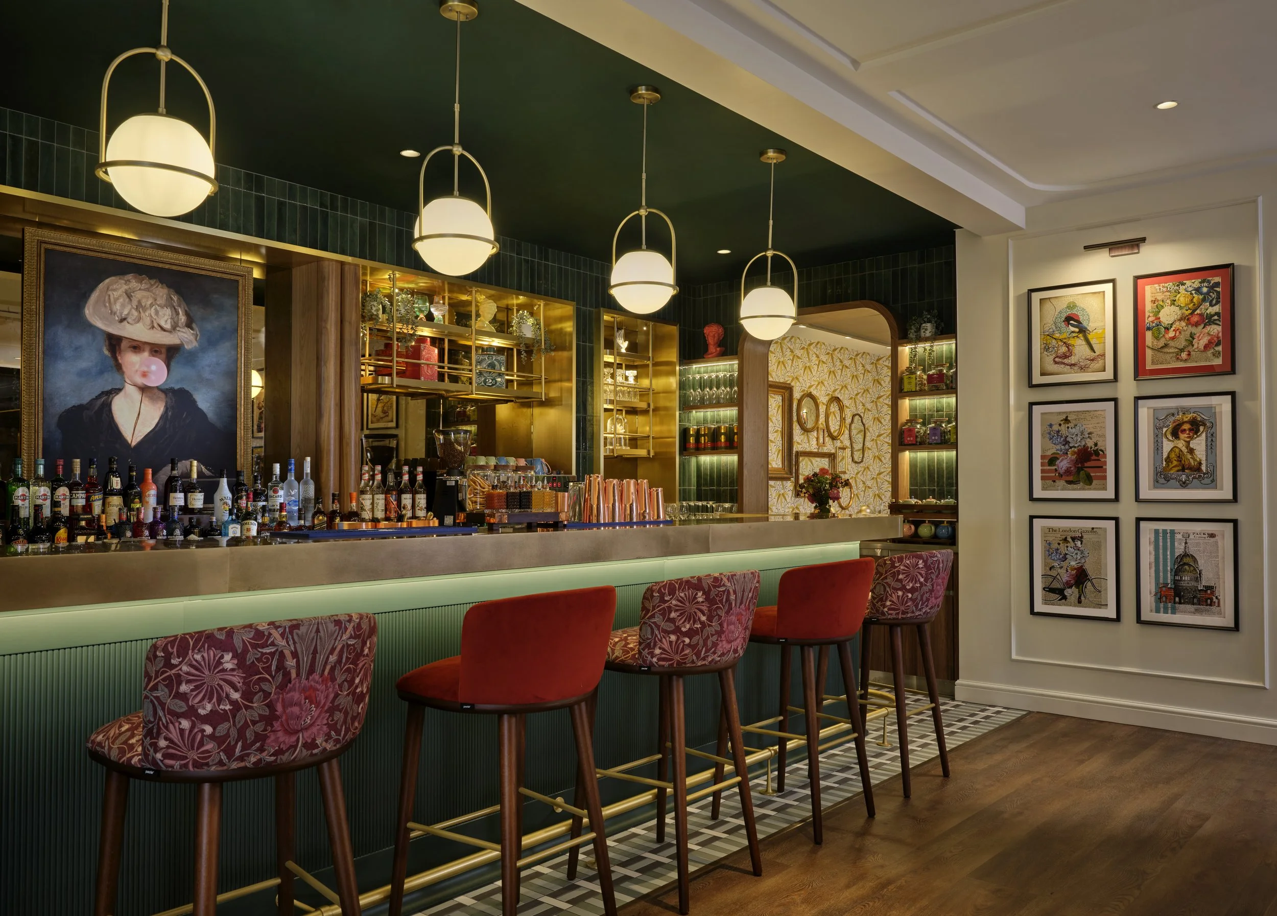 Interior of a bar with four hanging light fixtures, red bar stools with patterned upholstery, colorful framed artwork on the walls, shelves with glassware and decor, and a marble counter with various bottles of alcohol.
