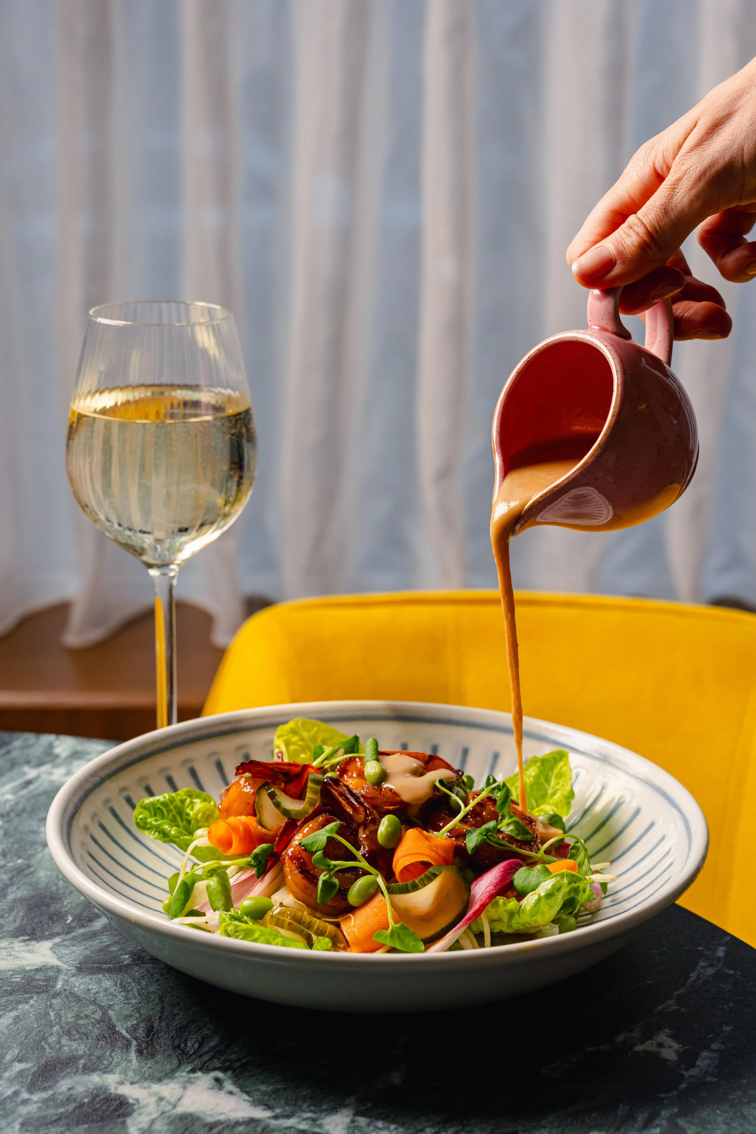 A bowl of mixed salad with vegetables and grilled meat on a dark marble table, a glass of white wine and a pink cup pouring sauce onto the salad, with a yellow chair in the background.