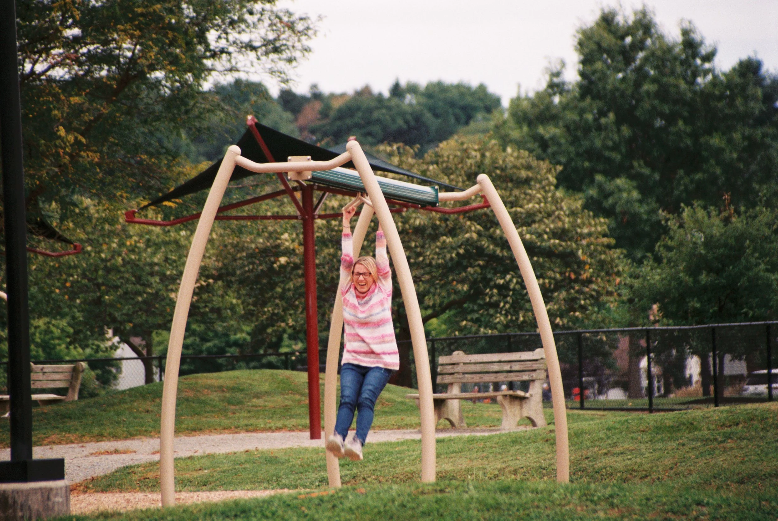 A girl with glasses and a pink and white striped sweater is swinging on a playground swing, smiling and looking happy, with trees and park benches in the background.