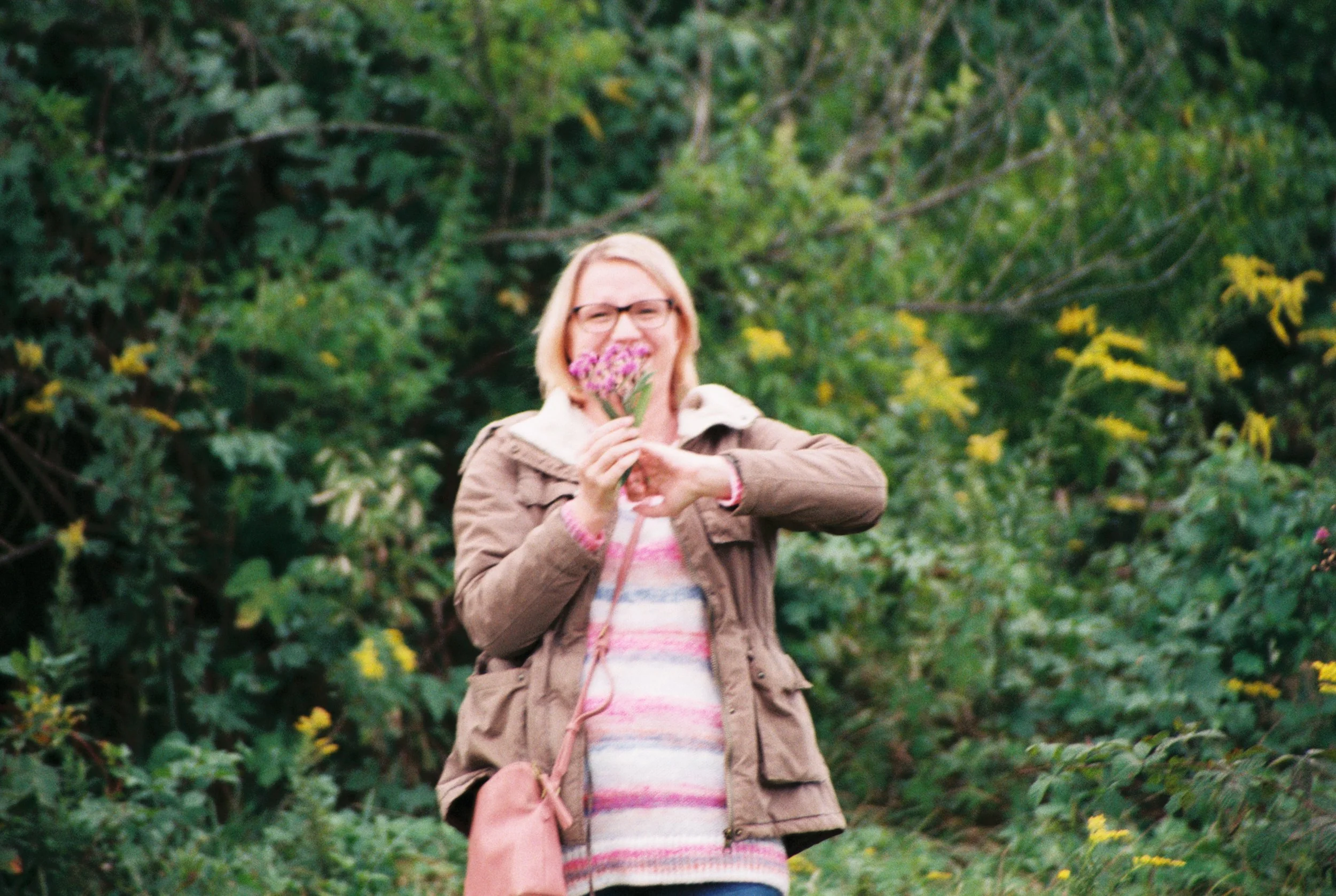 A woman with blonde hair and glasses holding a pink flower in front of her face, smiling, standing outdoors amidst green foliage.