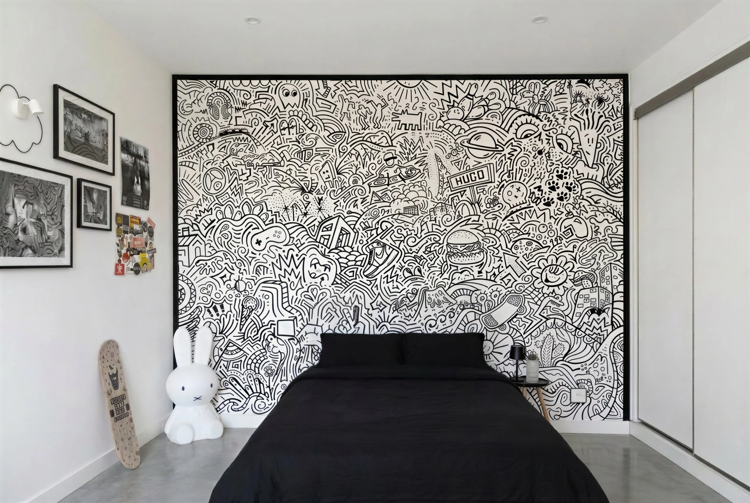 Mur noir et blanc avec des dessins abstraits et urbains, un canapé noir, une lampe de chevet noire, une figurine en forme de lapin blanc, un skateboard posé contre le mur, et des tableaux noirs et blancs accrochés au mur.