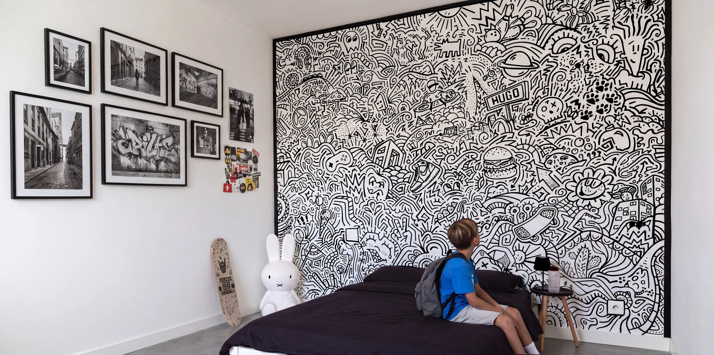 Chambre avec fresque doodle art graffiti noir et blanc, fresque personnalisée peinte à main levée, photos encadrées sur mur blanc, skateboard en bois, jeune garçon assis sur le lit bleu, bouteille d'eau sur table, décor moderne.