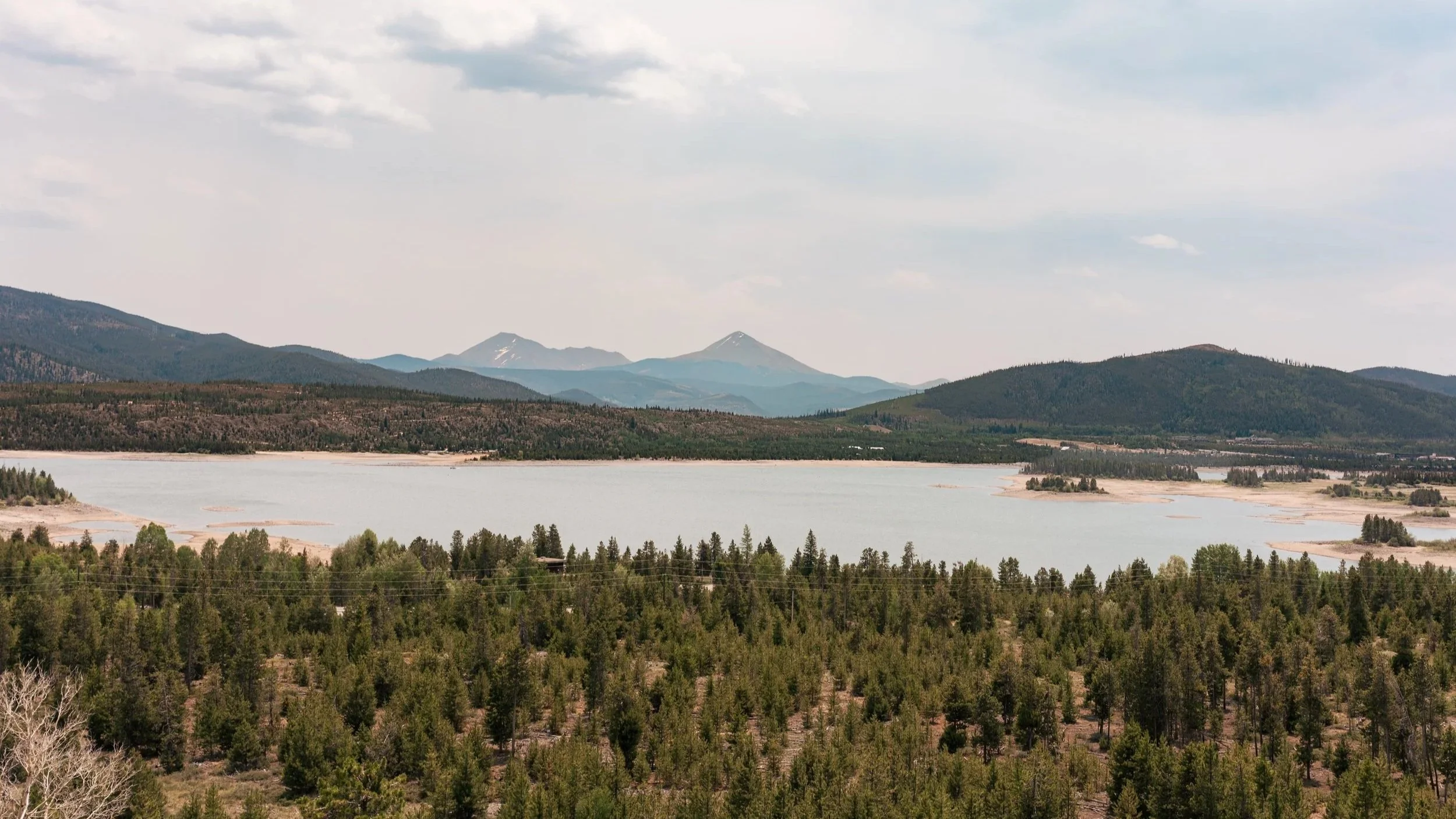A large body of water surrounded by green forests and mountains in the distance with snow-capped peaks