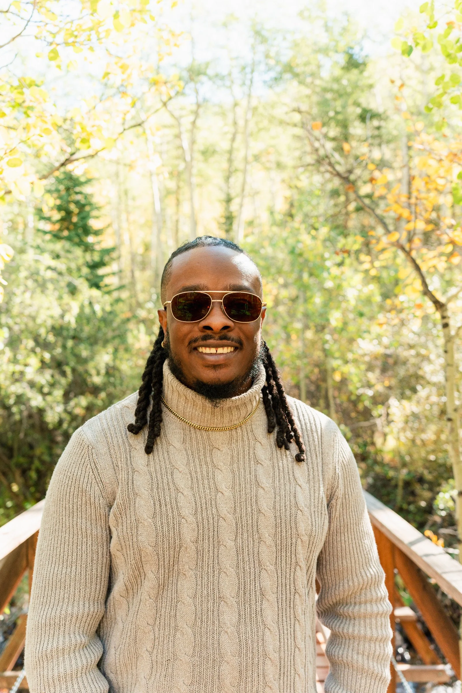 A man with dreadlocks wearing sunglasses and a knit sweater smiling outdoors on a wooden bridge in a forest with yellow and green trees.