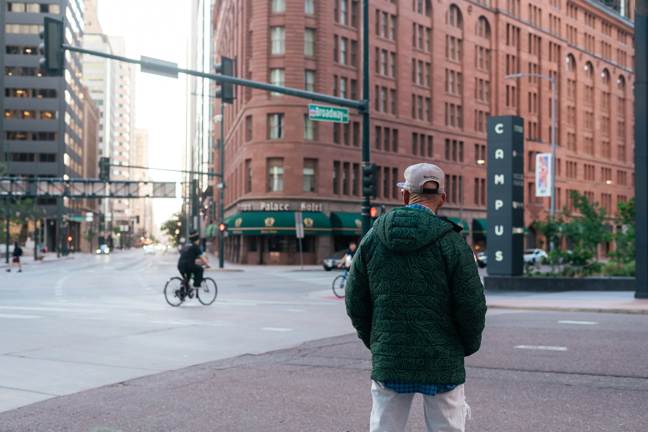 A person, seen from behind, wearing a light gray cap with '#staytrn' written on it, a green jacket, and light-colored pants, stands in an urban city intersection with tall red brick buildings, traffic lights, and streets with pedestrians and cyclists.