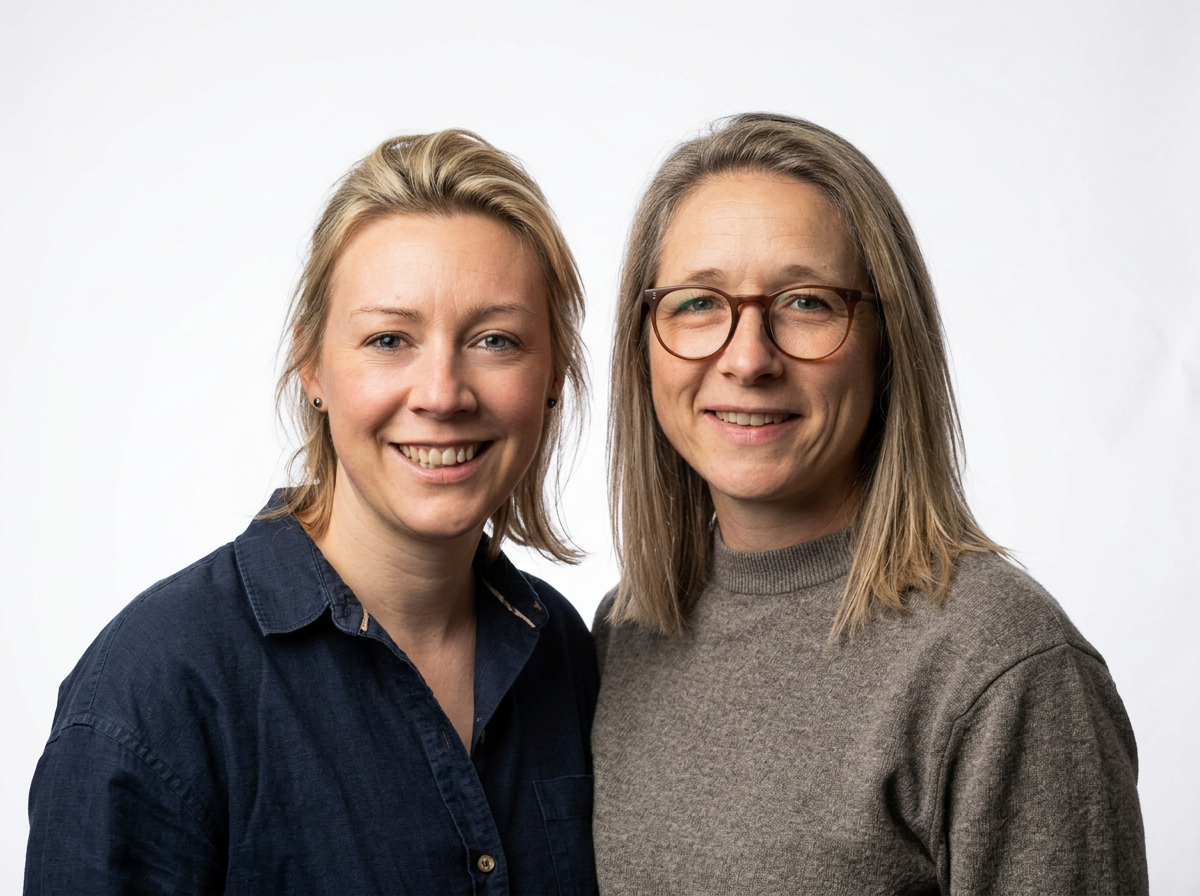 Two women smiling and standing close together against a plain white background, one with short blonde hair wearing a dark button-up shirt and the other with shoulder-length gray hair wearing glasses and a gray sweater.