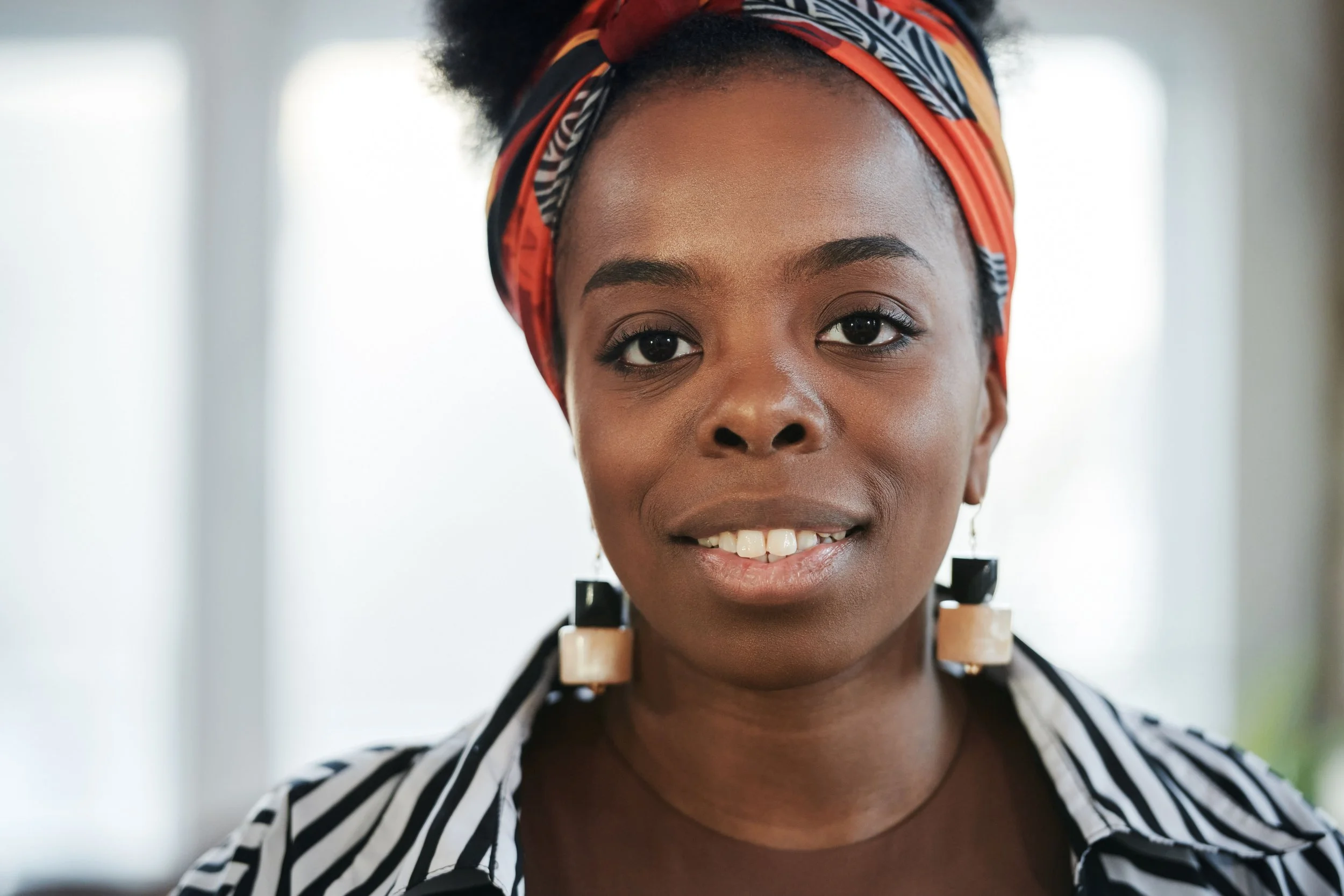Close-up of a smiling woman with dark skin wearing a colorful headscarf and earrings, with a blurred background.
