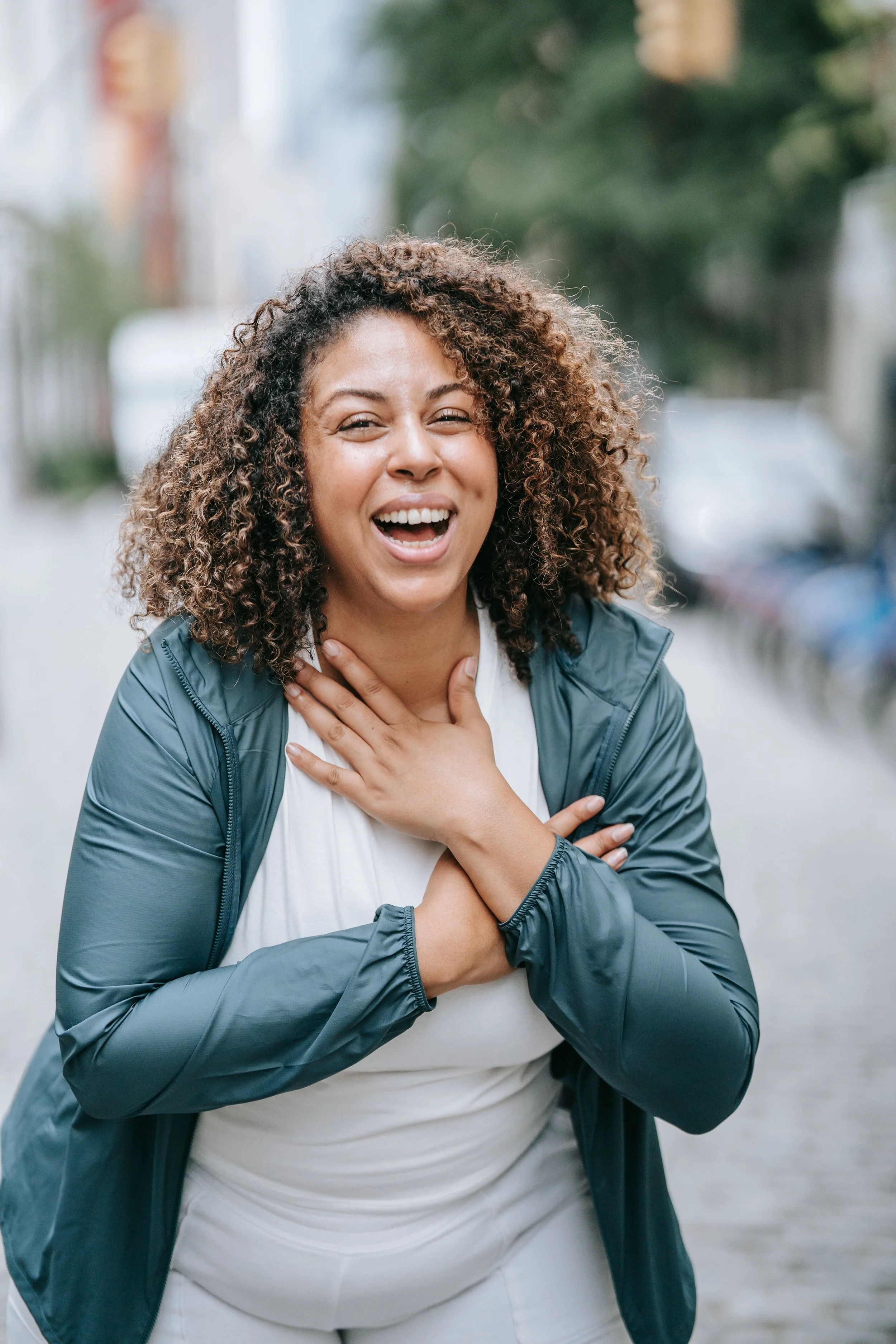 A woman with curly hair smiling outdoors, wearing a gray jacket and white shirt, with her hand on her chest.