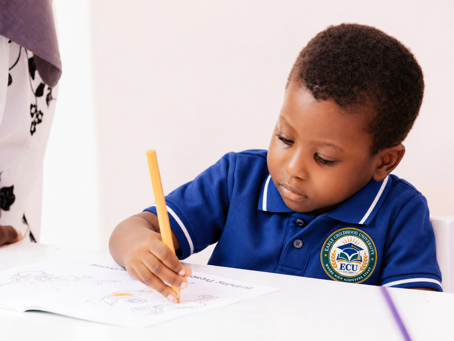 A young boy wearing a blue polo shirt with an ECU logo is drawing with a yellow pencil at a white table.