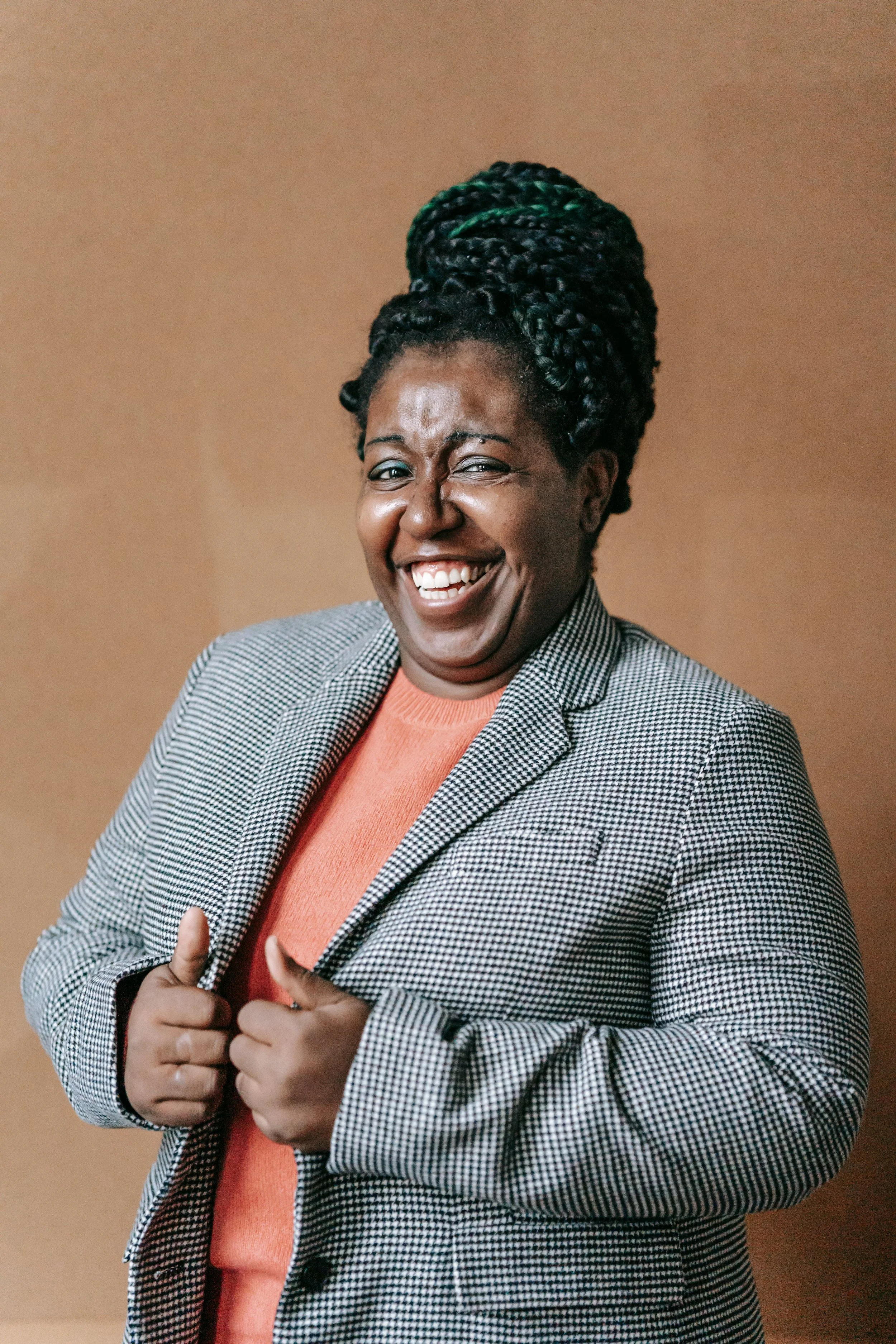 A woman with braided hair in a high bun, wearing a checkered blazer and an orange top, smiling and giving two thumbs up.