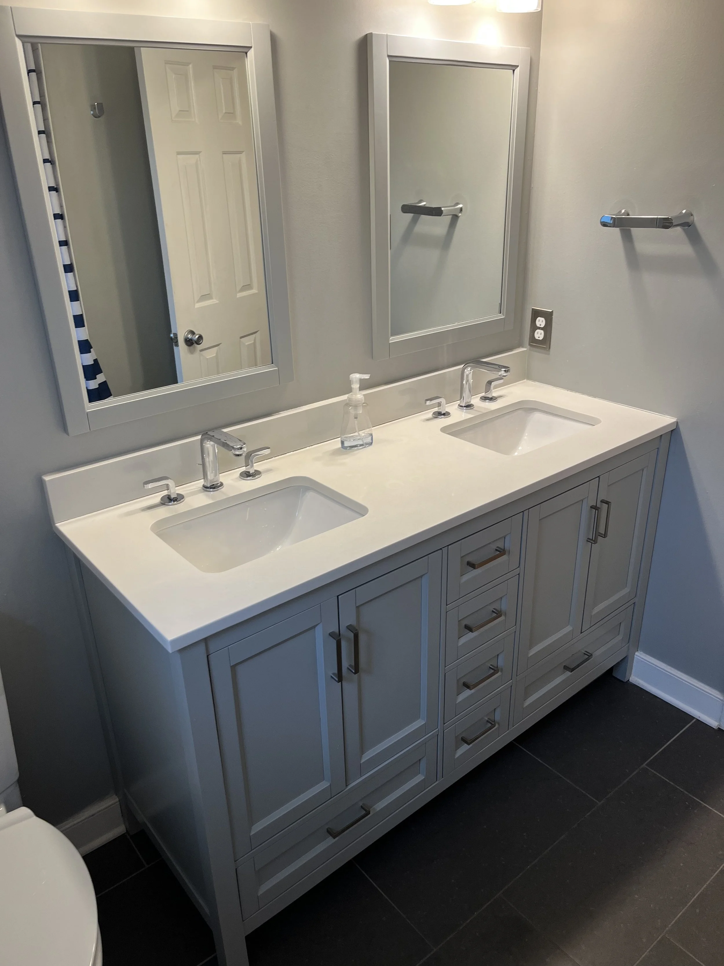A modern double bathroom vanity with a white countertop, two sinks, and two mirrors with silver frames. A bottle of hand soap is placed between the sinks, and the vanity has multiple drawers and cabinets with silver handles. The room has gray walls, a white door, and dark floor tiles.