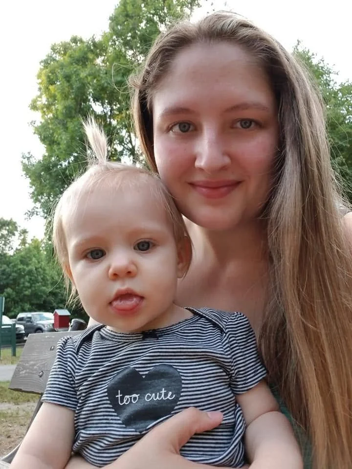 A woman and a young girl taking a selfie outdoors. The girl is wearing a striped shirt with a black heart and the words 'too cute' on it. Trees and parked cars are visible in the background.
