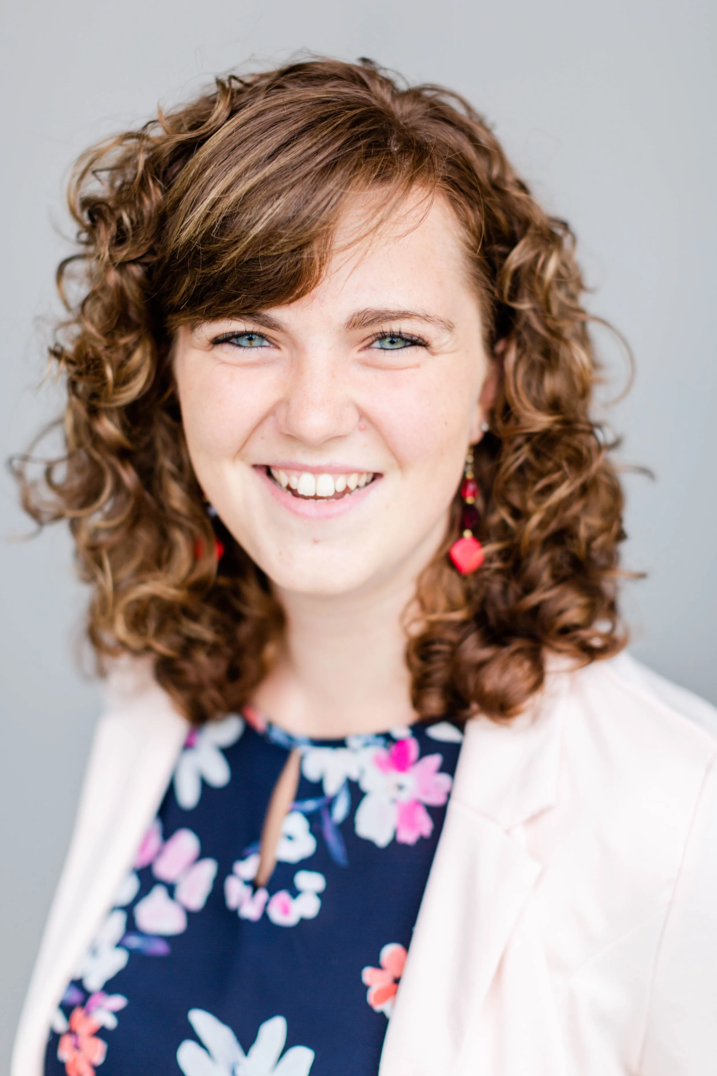 Close-up portrait of a woman with curly reddish-brown hair, blue eyes, smiling, wearing colorful earrings, floral dress, and a light-colored blazer against a neutral background.