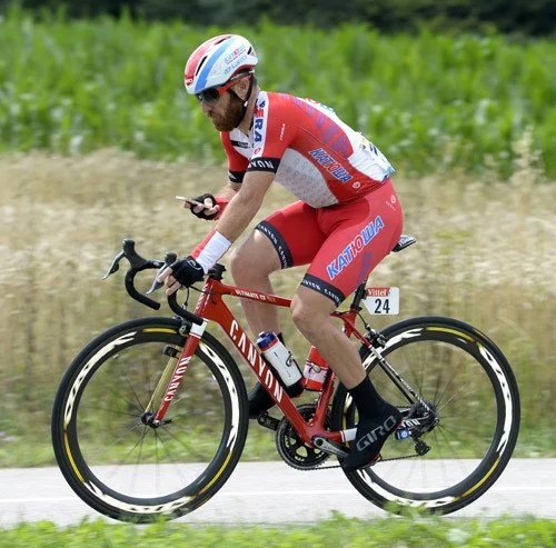 Cyclist in red and white racing uniform riding a red Canyon bike on a rural road, looking at a phone with green fields in the background.