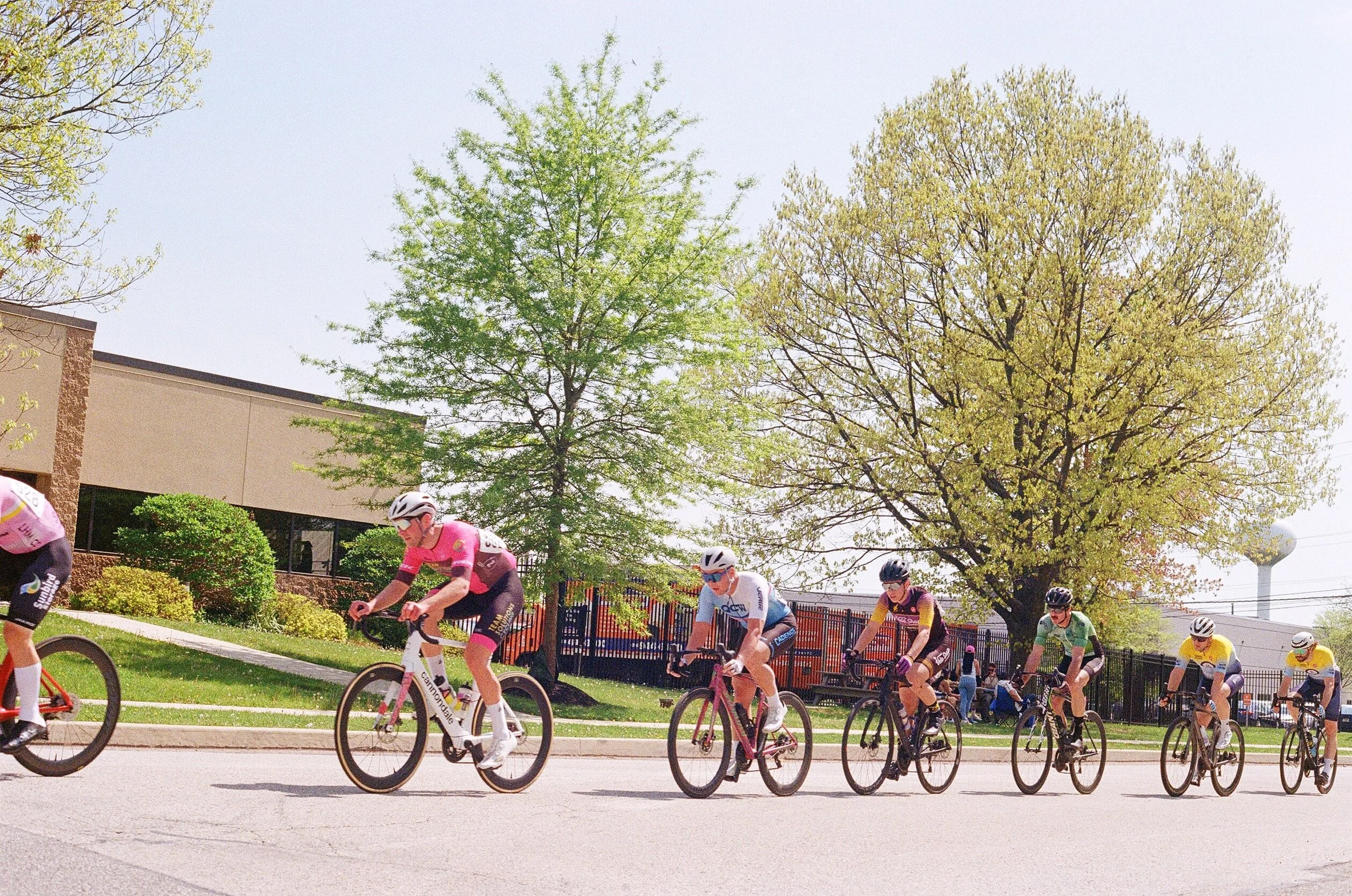 A group of cyclists riding on a street with green trees and a building in the background during daytime.